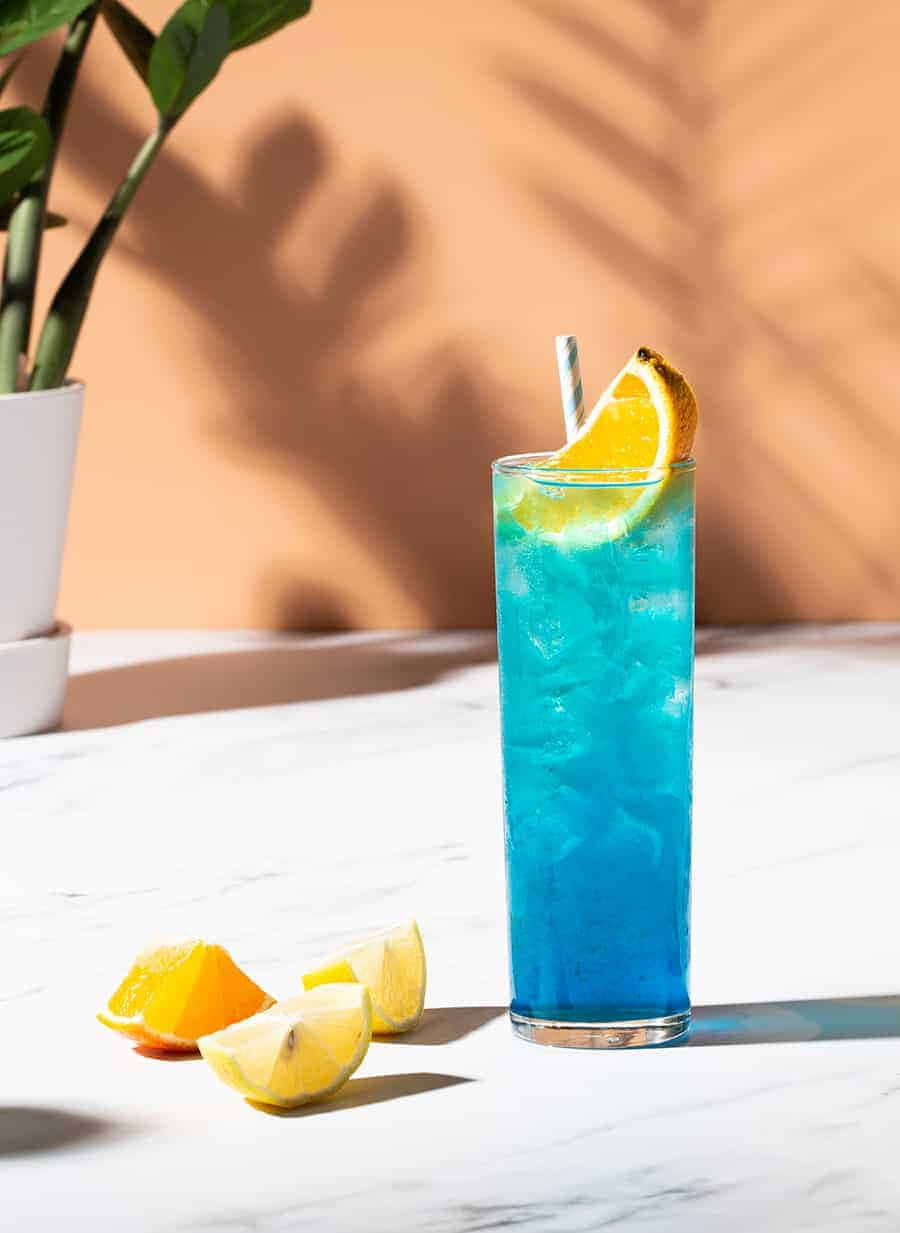 Beautiful Drink 桌布 壁紙 — 一杯充滿活力的藍色雞尾酒，加冰和柳橙片，放在大理石桌上，旁邊有柑橘楔形塊，沐浴在溫暖的陽光下。