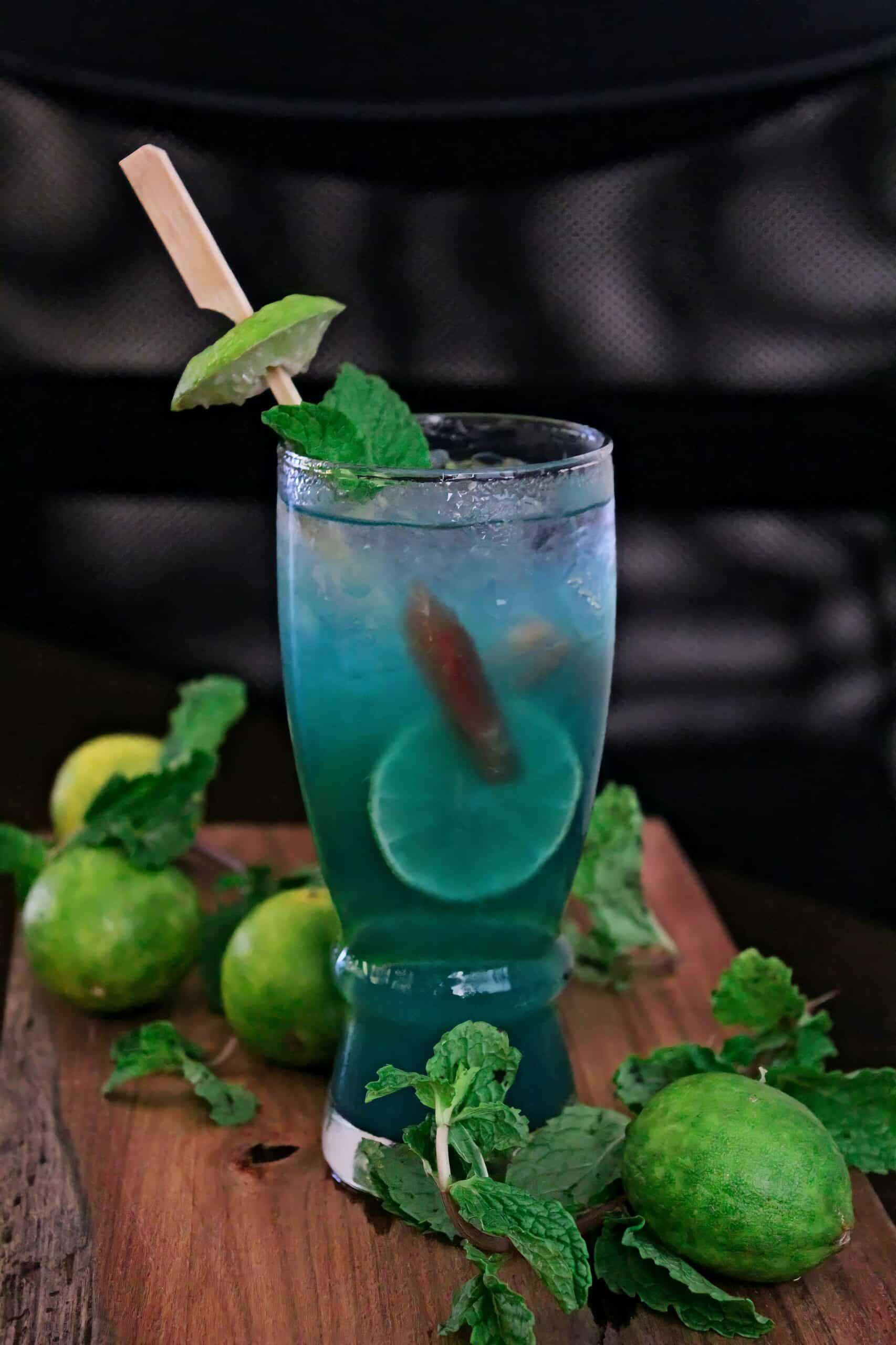 Beautiful Drink 桌布 壁紙 — 一杯充滿活力的藍色雞尾酒，飾以萊姆和薄荷，放在木板上，周圍是新鮮的萊姆和薄荷葉。