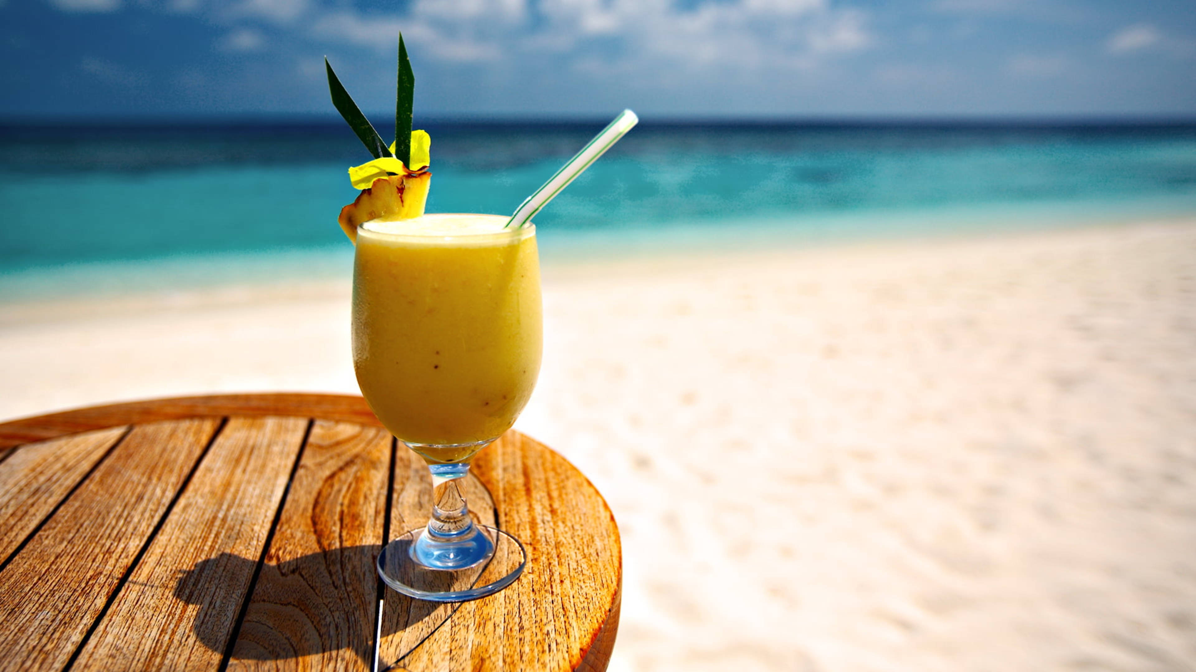 Tropical Drink 桌布 壁紙 — 一杯充滿活力的黃色熱帶飲料，配上鳳梨裝飾，放在木桌上，背景是藍綠色的海洋和白色沙灘。