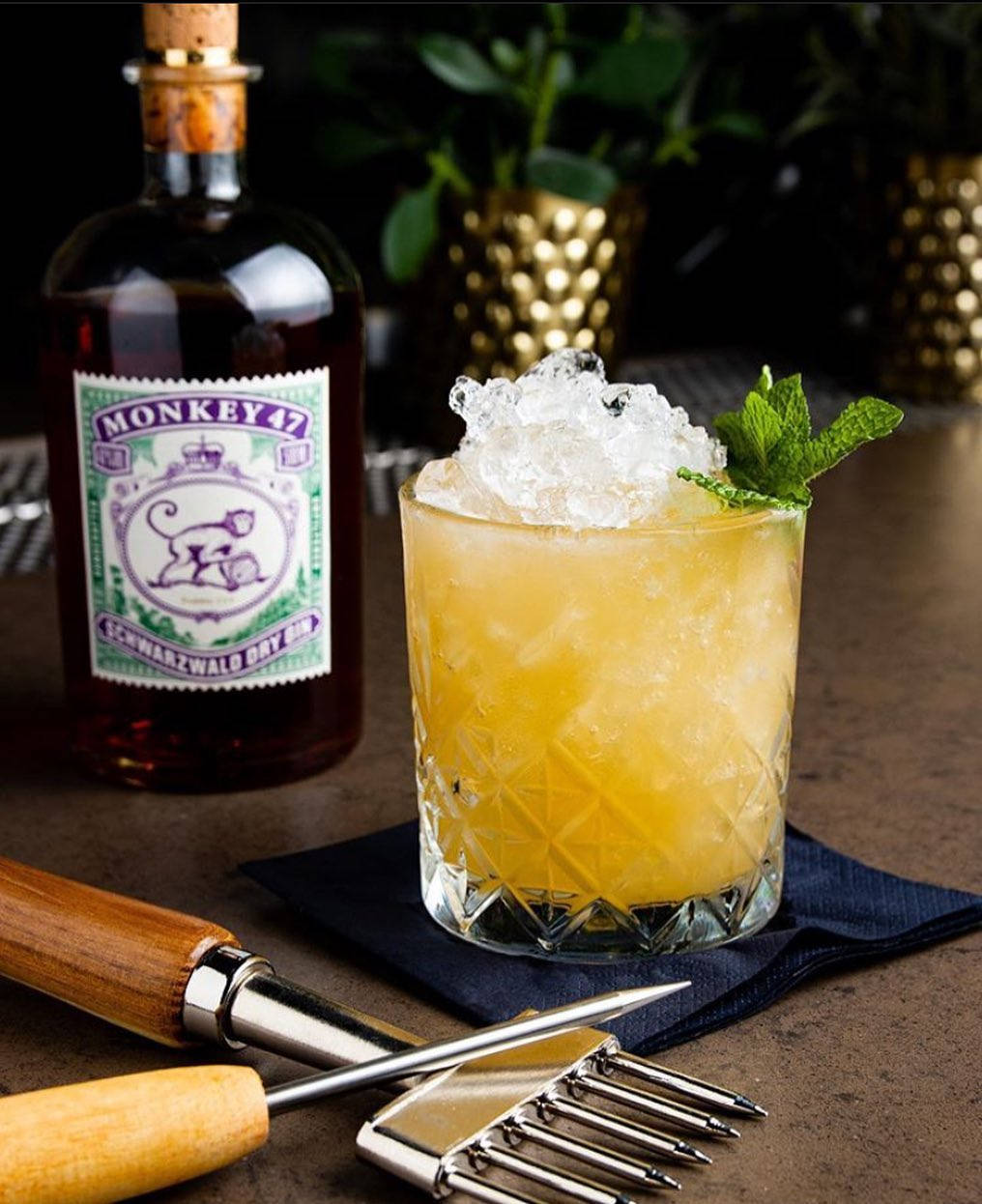 cocktail 桌布 壁紙 — 一杯充滿活力的黃色雞尾酒，配上碎冰和薄荷裝飾，旁邊放著一瓶Monkey 47琴酒和酒吧工具，置於深色餐巾上。