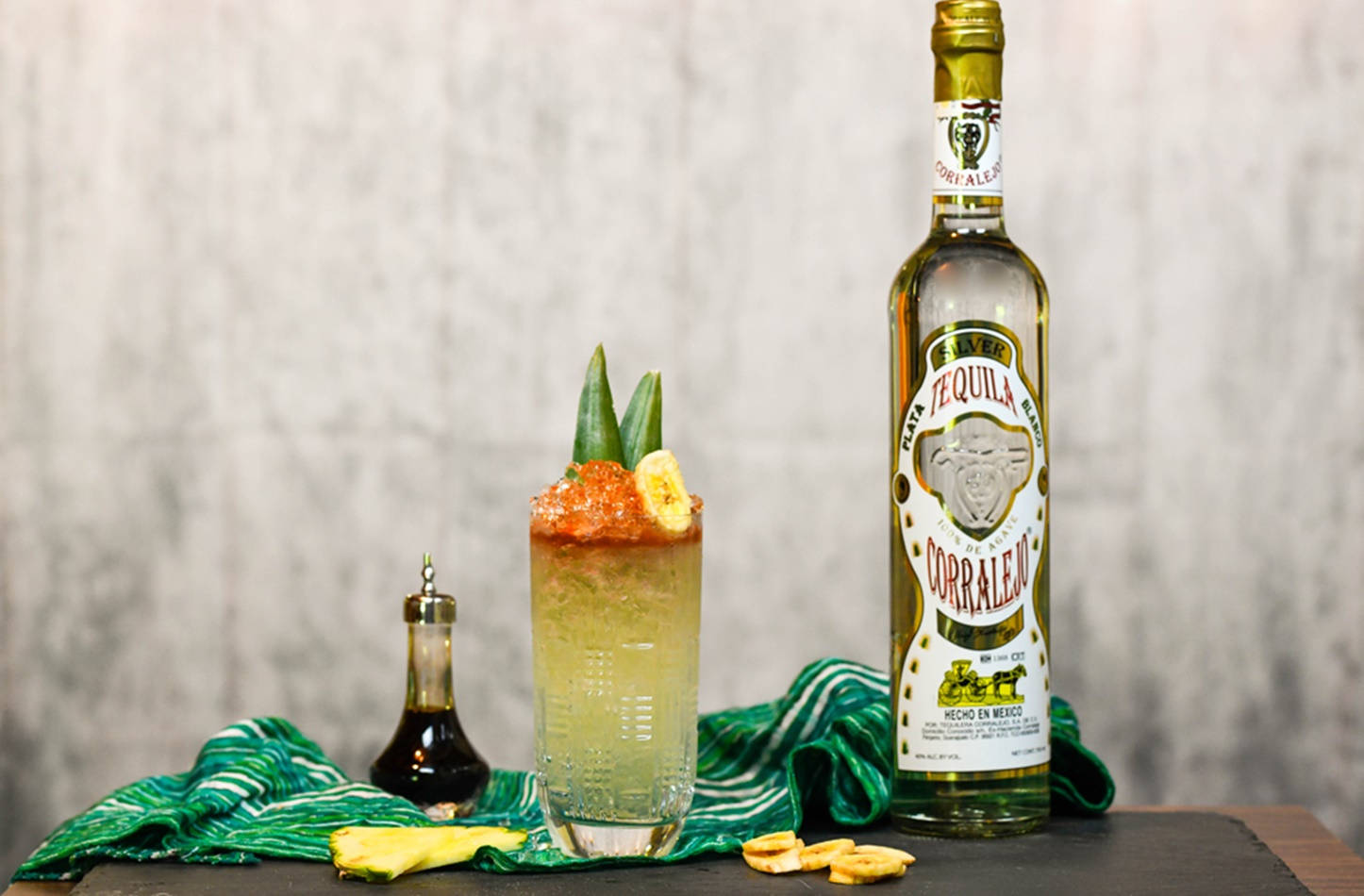 cocktail 桌布 壁紙 — 一杯充滿活力的龍舌蘭雞尾酒，以香蕉和龍舌蘭裝飾，旁邊放著一瓶Corralejo龍舌蘭酒，置於綠色布料上，背景為紋理牆壁。