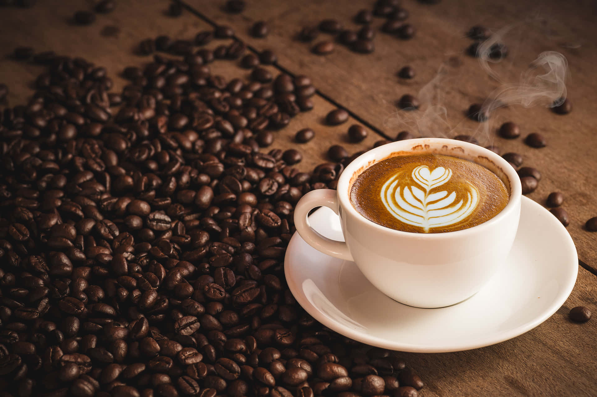 Coffee Cup 桌布 壁紙 — 一杯冒煙的咖啡，帶有心形拉花藝術，放在木桌上，周圍散落著深烘焙咖啡豆。