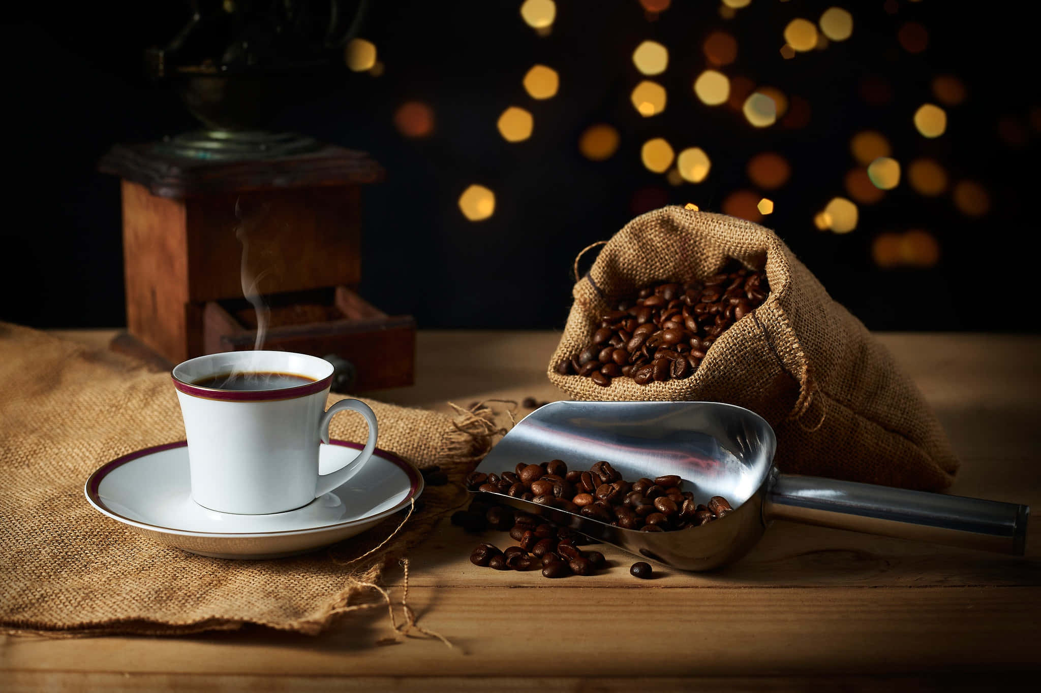 Coffee Aesthetic 桌布 壁紙 — 一杯冒煙的咖啡放在木桌上，旁邊有一個麻布袋和一個裝滿烘焙咖啡豆的勺子，背景有溫暖的散景燈光。