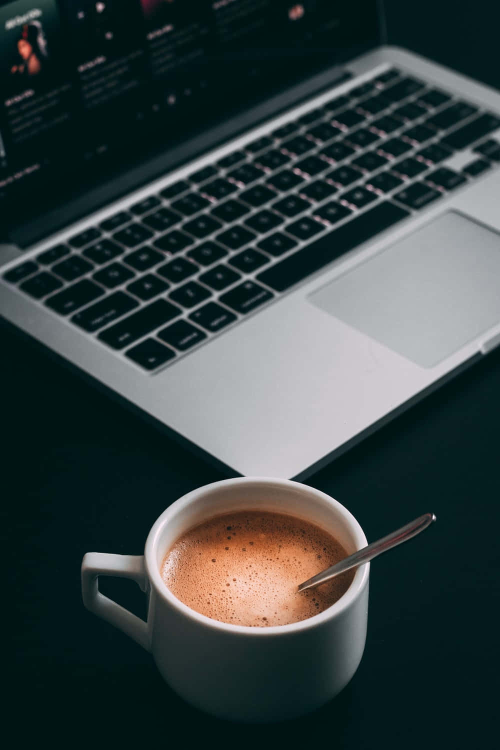Minimalist Coffee 桌布 壁紙 — 一杯冒煙的咖啡配上一隻湯匙，放在昏暗的桌子上，旁邊是一部筆記型電腦，在微光中柔和地發光。