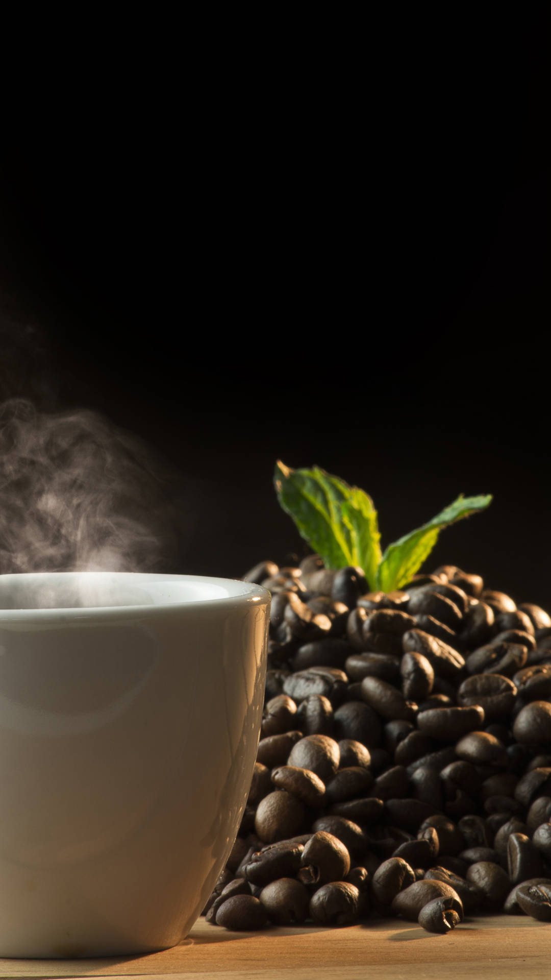 Coffee Aesthetic 桌布 壁紙 — 一杯冒煙的白色咖啡杯，旁邊有一堆烘焙過的咖啡豆和新鮮薄荷葉，背景深色。