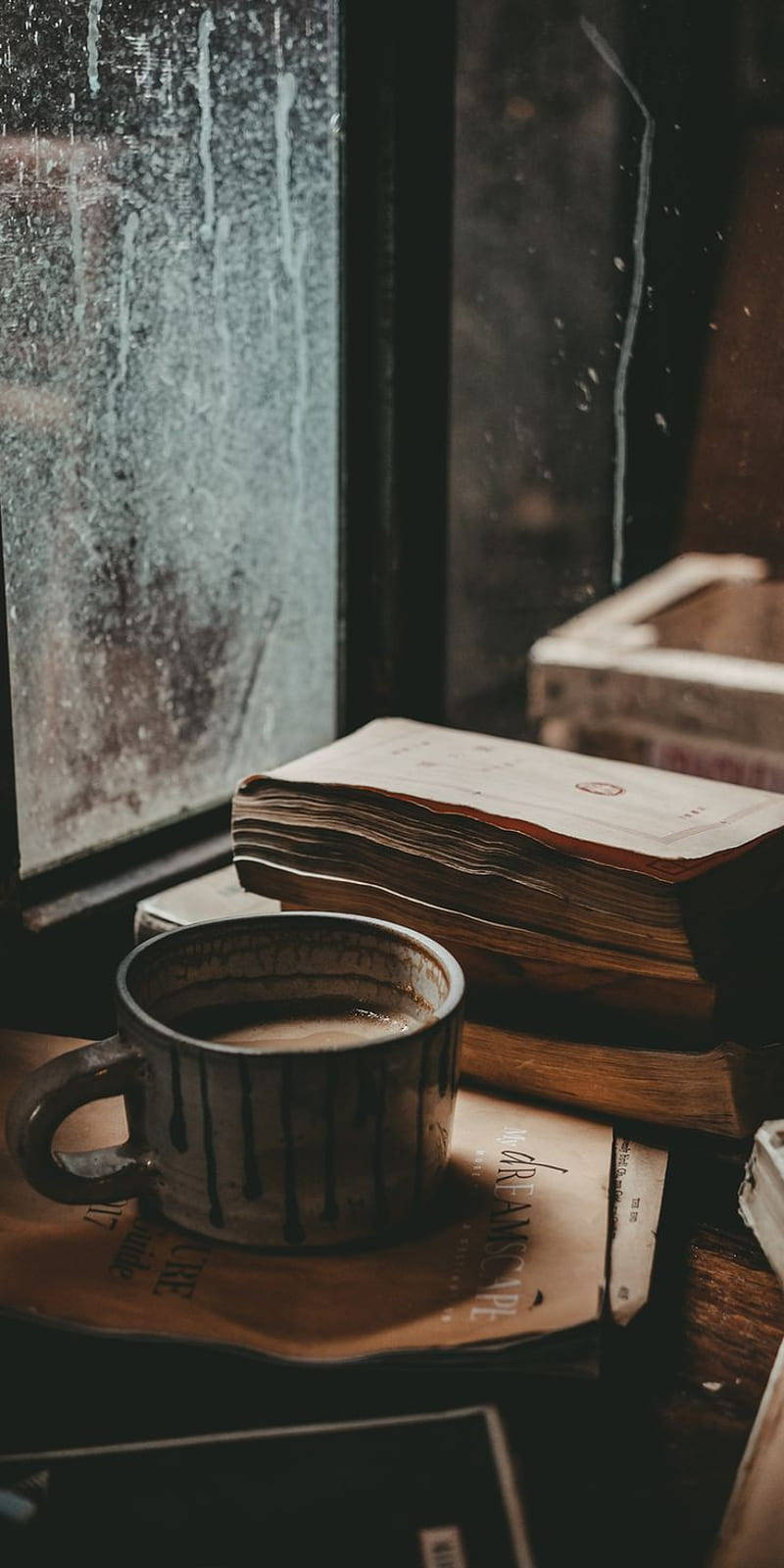 咖啡動圖 桌布 壁紙 — 一杯冒煙的馬克杯放在一堆書上，旁邊是雨水斑駁的窗戶，在一個舒適昏暗的房間裡。