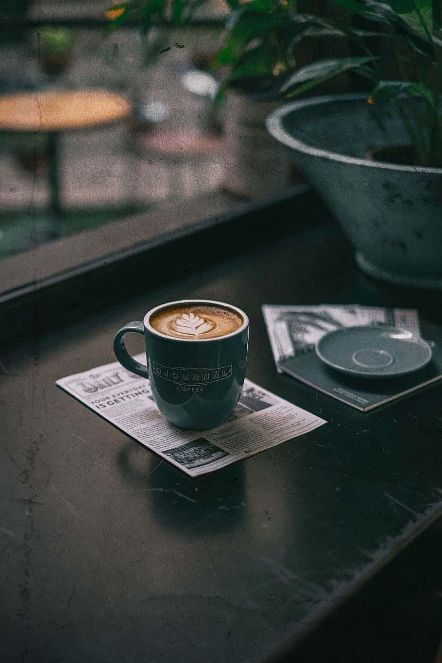 Coffee Aesthetic 桌布 壁紙 — 一杯冒著熱氣的拉花咖啡放在報紙上，置於深色桌子，旁邊有一盆盆栽，營造出舒適的咖啡館氛圍。