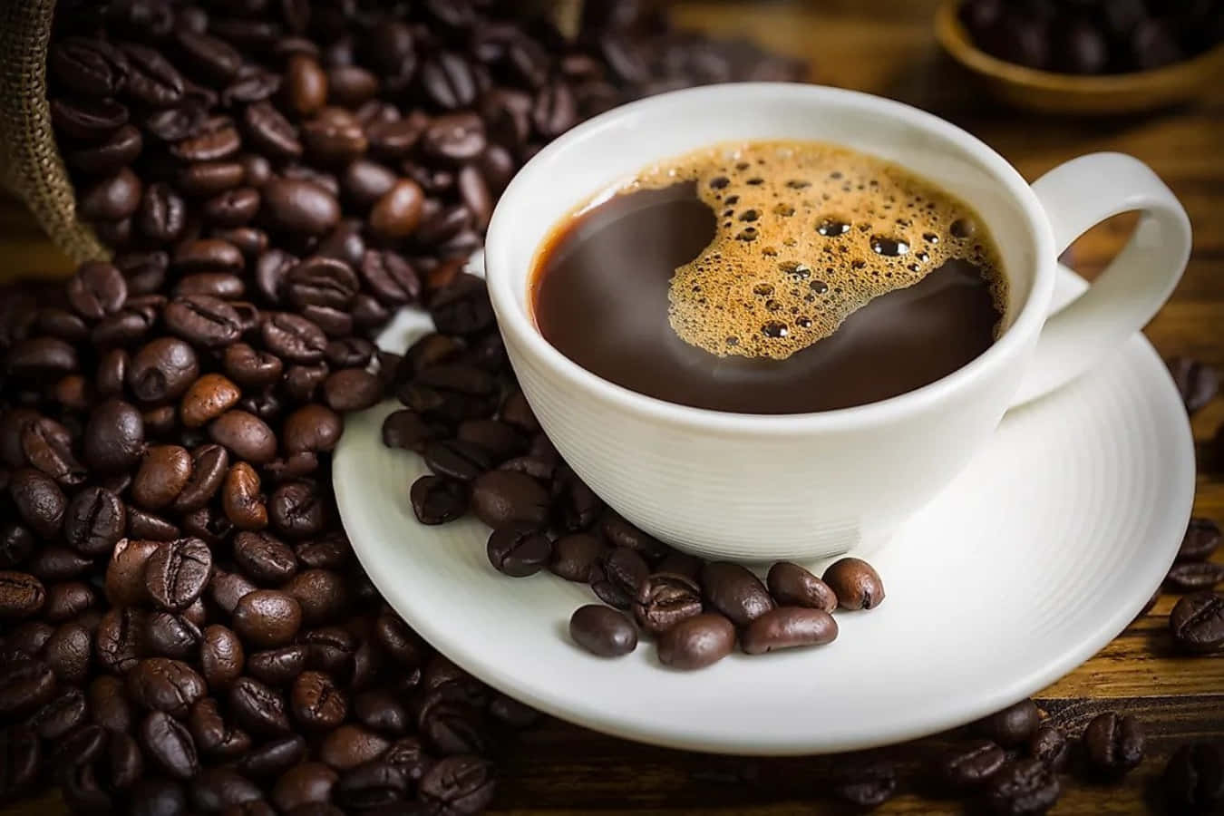 Coffee Cup 桌布 壁紙 — 一杯冒著熱氣的白咖啡，帶有豐富的奶泡，被烘焙咖啡豆包圍，放在質樸的木桌上。