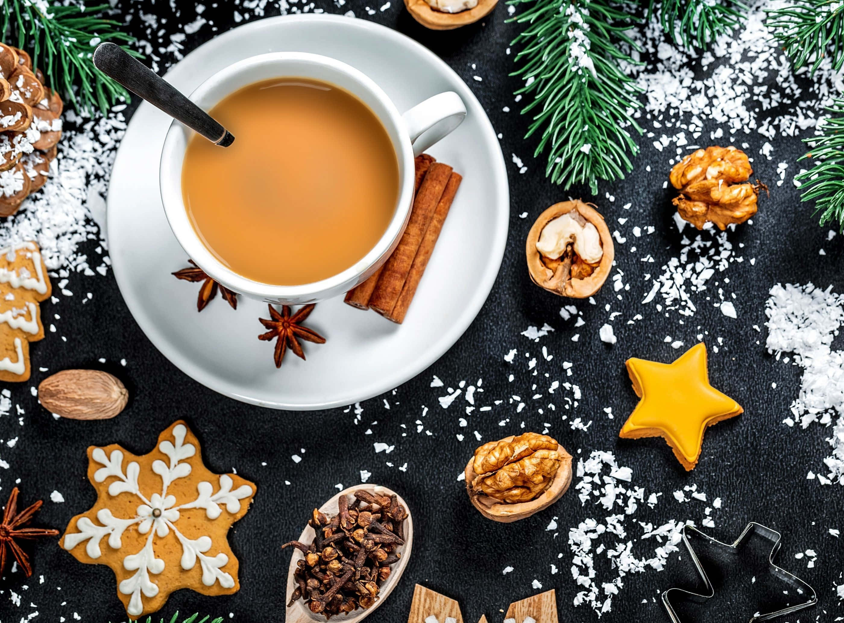 Winter Food 桌布 壁紙 — 一杯冒著熱氣的香料茶，周圍環繞著節日薑餅、肉桂棒和松針，放在白雪覆蓋的黑色表面上。