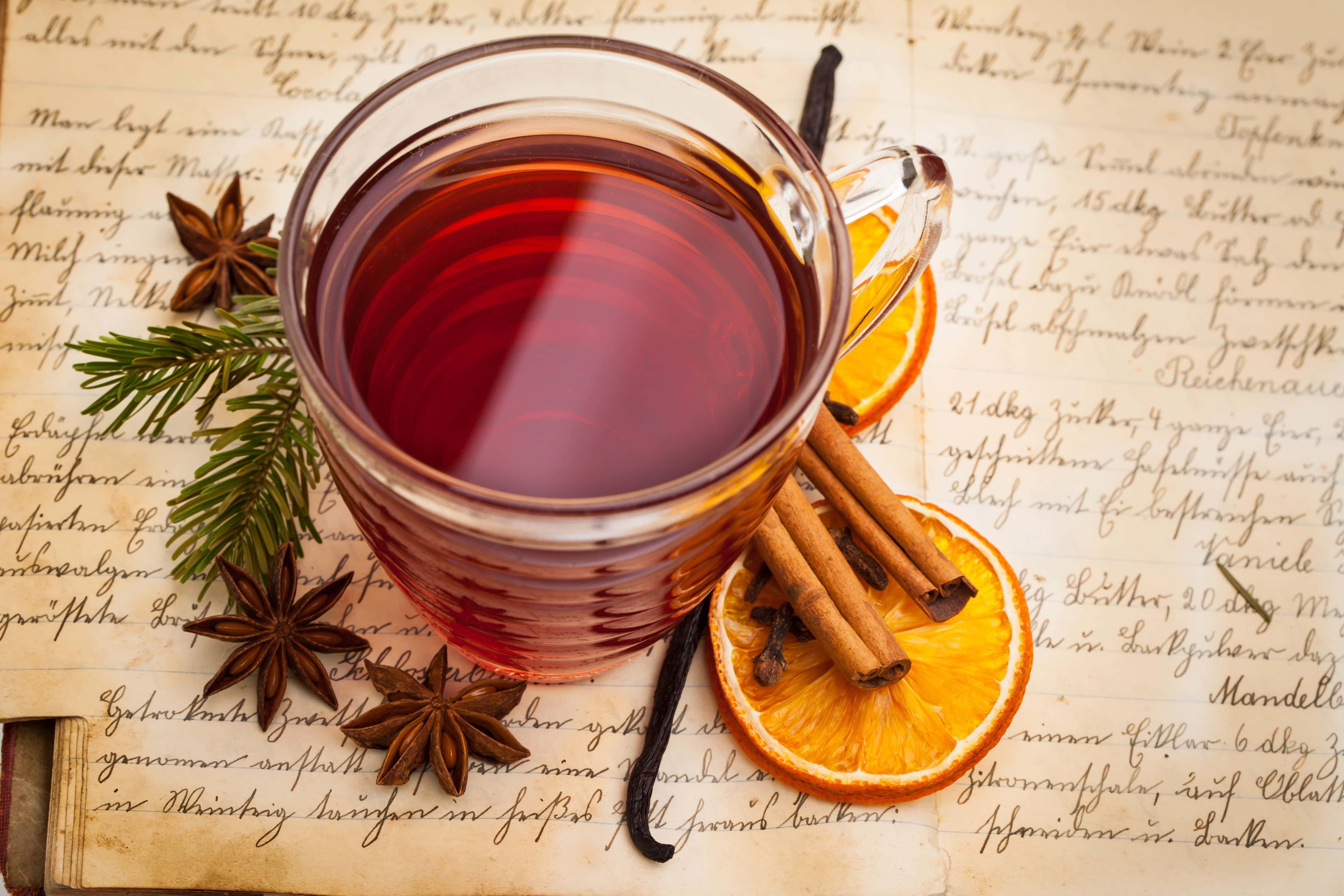 Mulled Wine 桌布 壁紙 — 一杯冒著蒸氣的香料茶玻璃杯放在復古手寫紙上，周圍有肉桂棒、八角、橙片和香草豆。