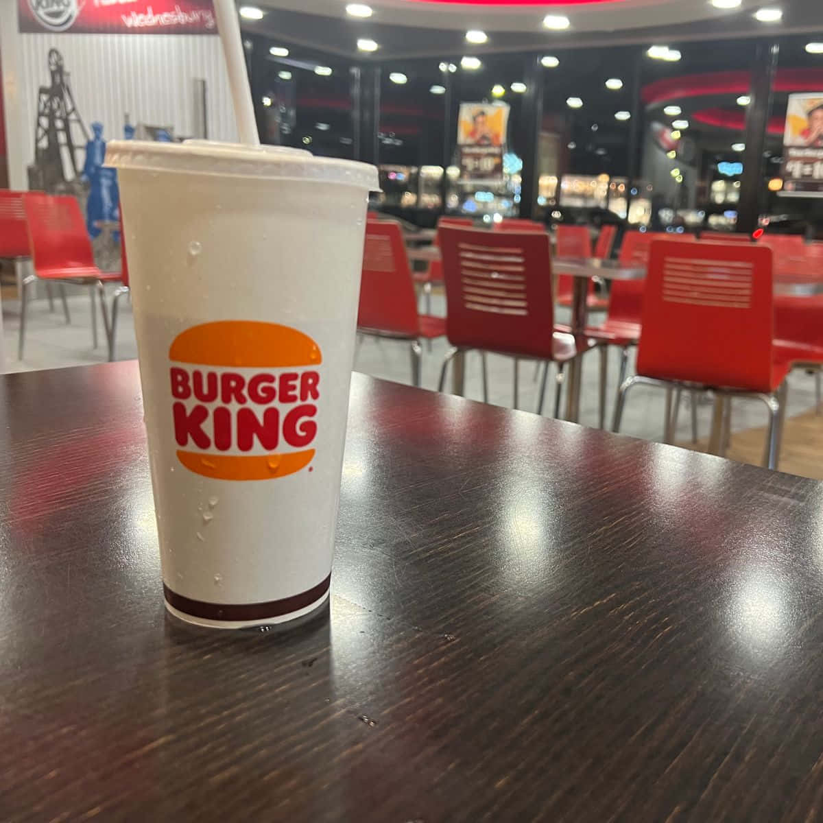 漢堡王 桌布 壁紙 — 一杯冰冷的漢堡王飲品，帶有冷凝水珠，放在夜間光線明亮的速食店的深色木桌上。