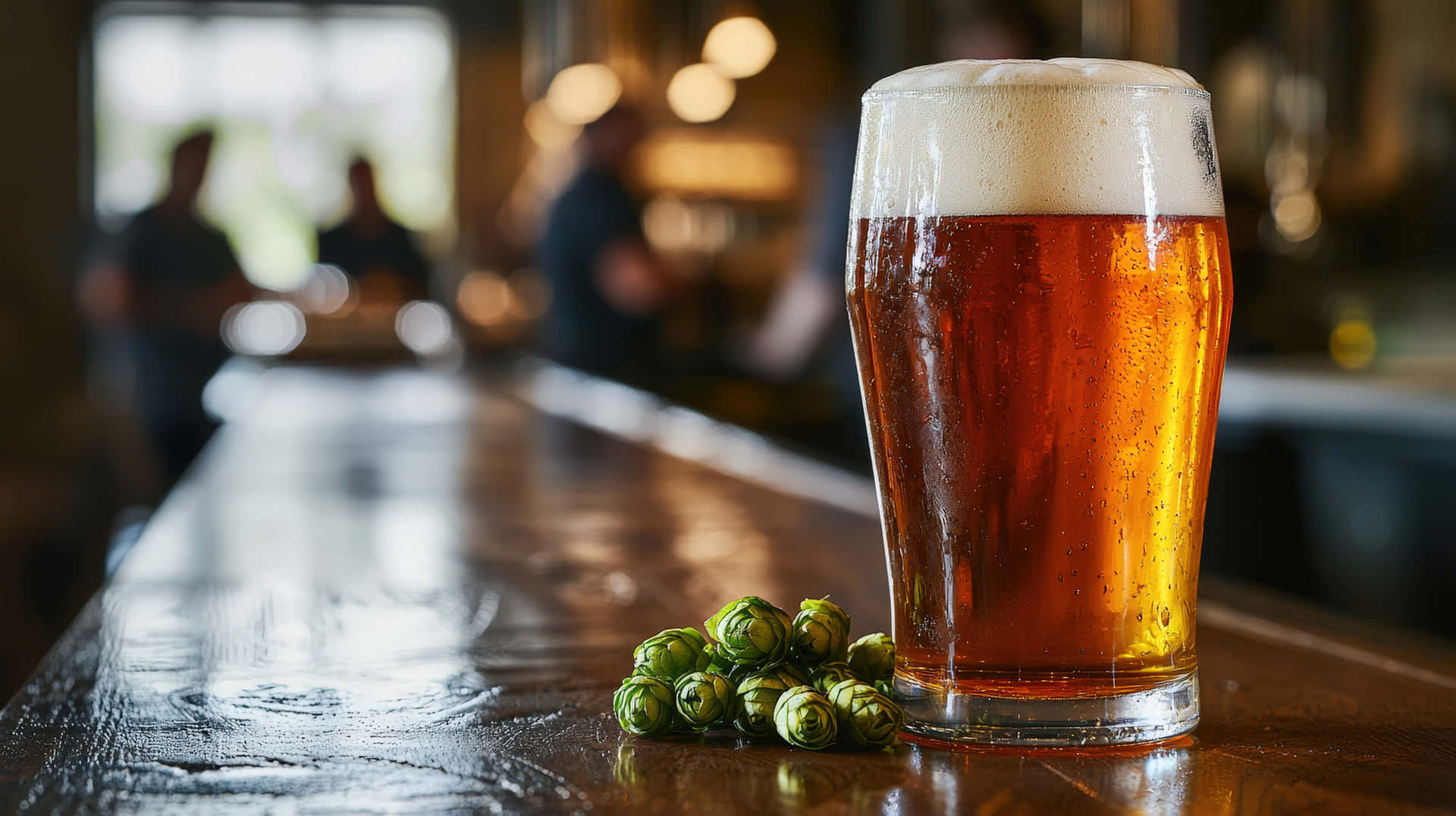 A frosty pint of amber beer sitting on a wooden bar counter in a cozy pub 壁紙 — 一杯冰凍琥珀色啤酒，帶有白色泡沫，放在拋光木吧檯上，旁邊是新鮮的綠色啤酒花球果。