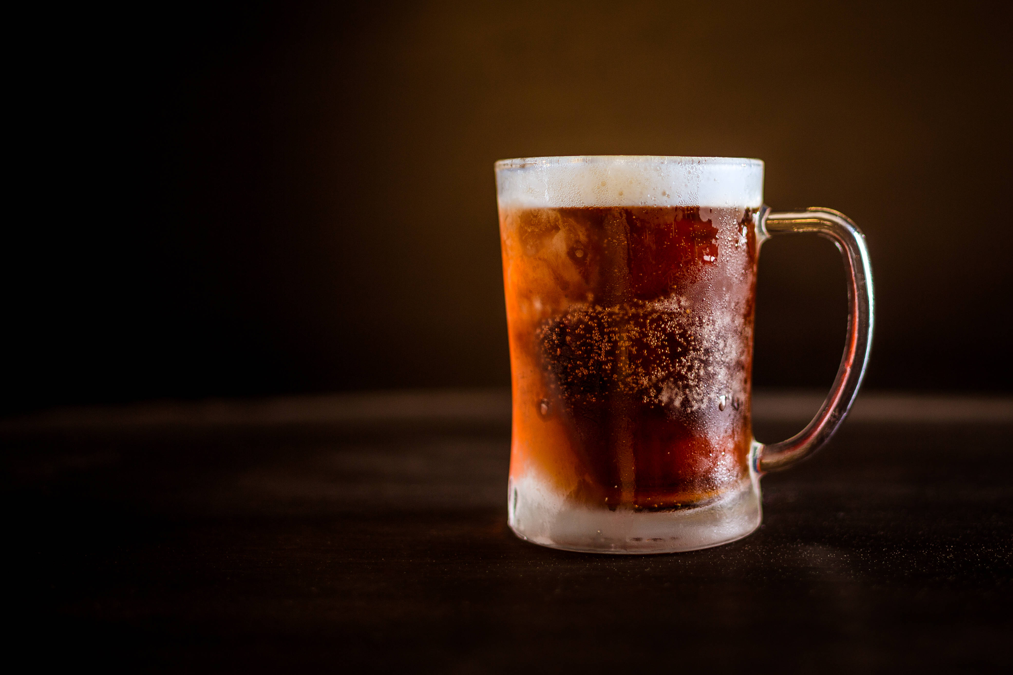 Beer 桌布 壁紙 — 一杯冰凍琥珀色啤酒杯，帶有泡沫和冷凝水，放在深色木質表面上，背景是溫暖的棕色。