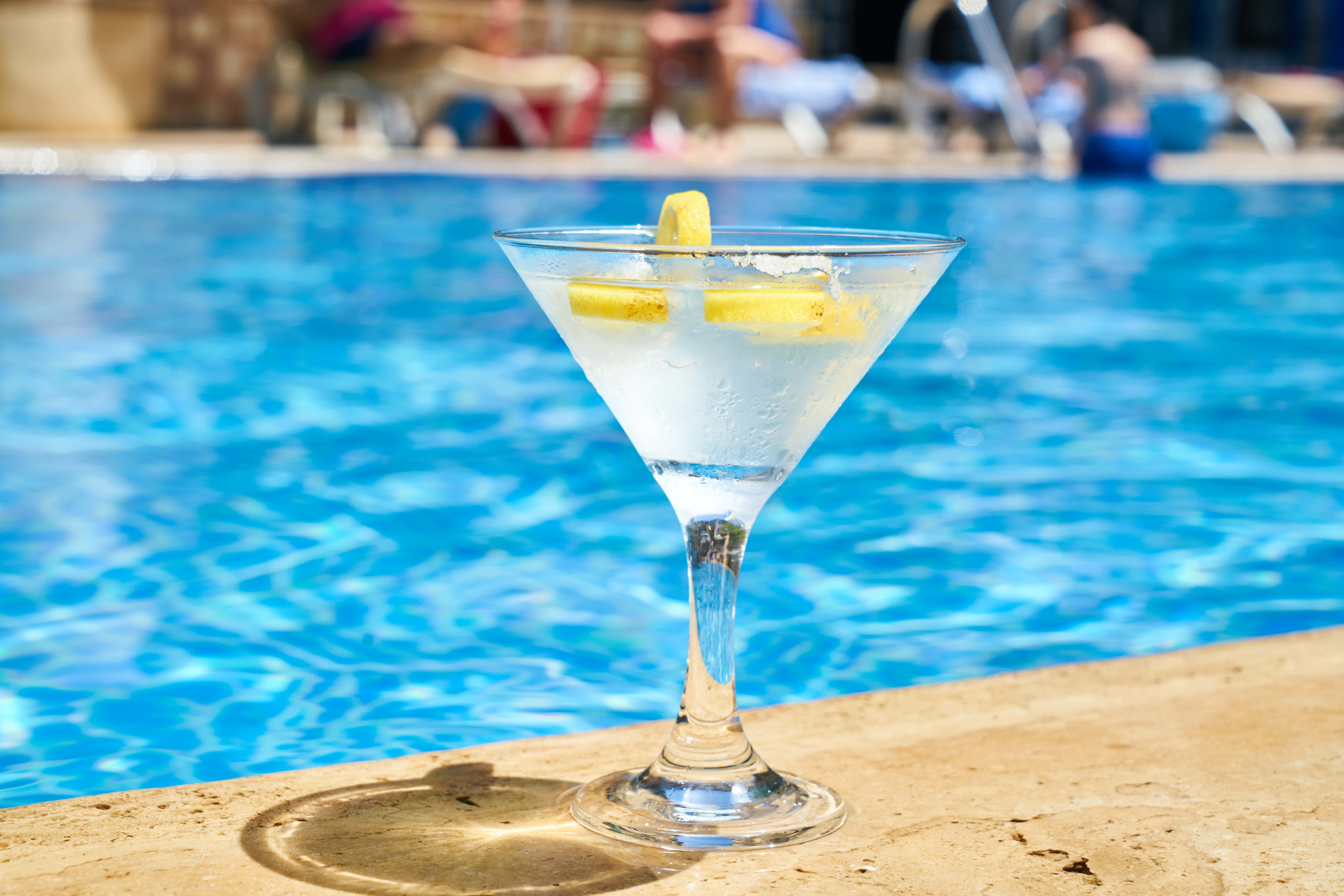 Summer Drinks 桌布 壁紙 — 一杯冰凍馬丁尼配檸檬裝飾，放在陽光普照下閃爍藍色水邊的泳池石上。