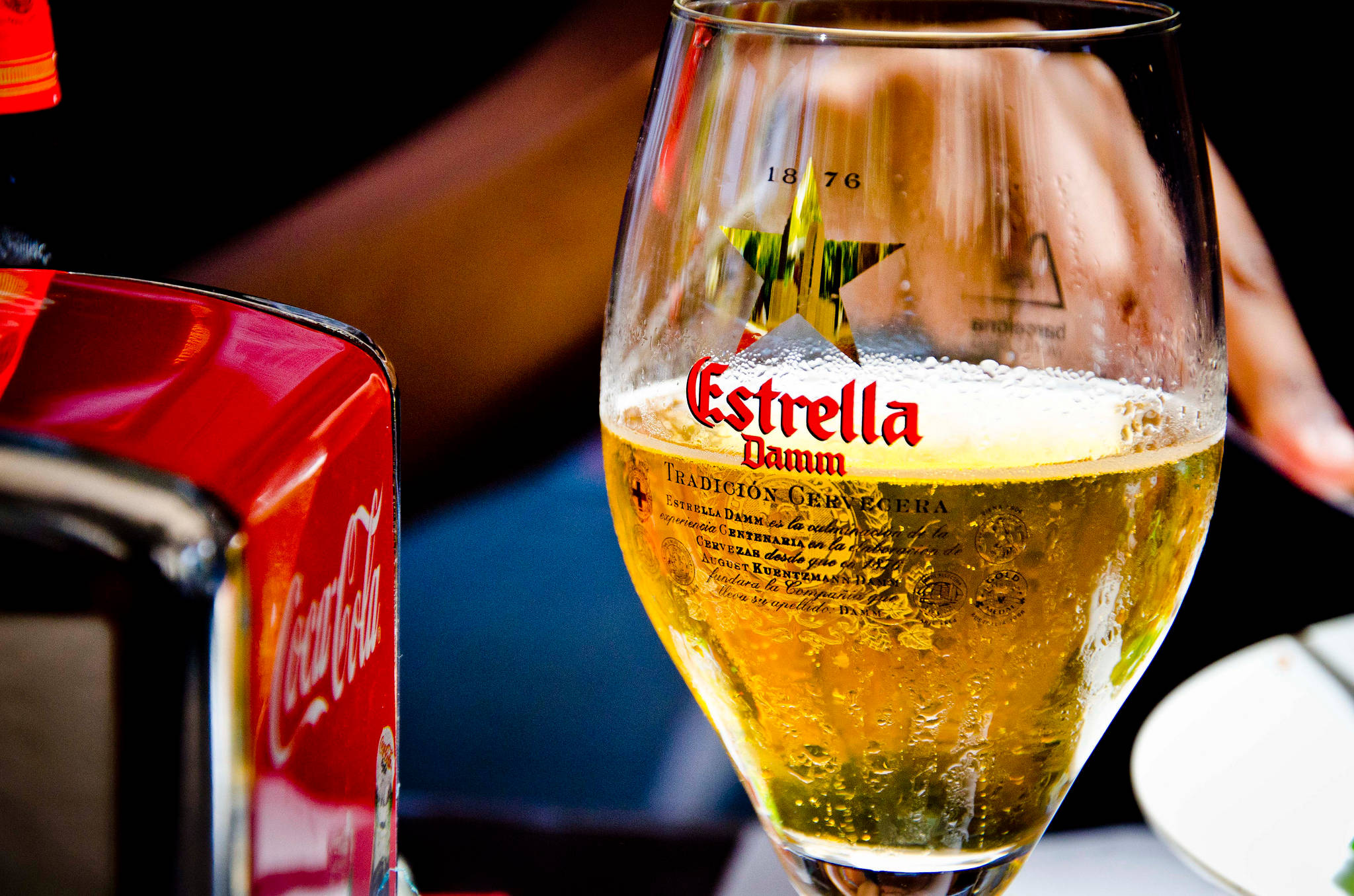 Drink 桌布 壁紙 — 一杯冰涼的 Estrella Damm 啤酒，帶有冷凝水珠，放在一瓶紅色可口可樂旁邊，環境燈光昏暗。