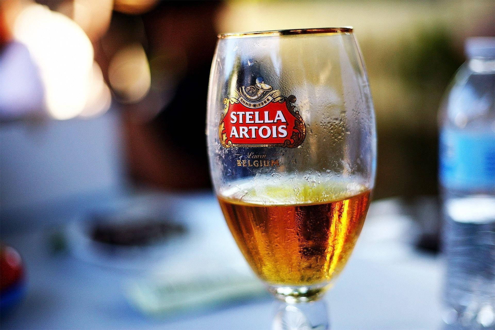 飲料 桌布 壁紙 — 一杯冰涼的 Stella Artois 啤酒杯，杯身凝結水珠，放在戶外桌子上，沐浴在溫暖的金色陽光中。