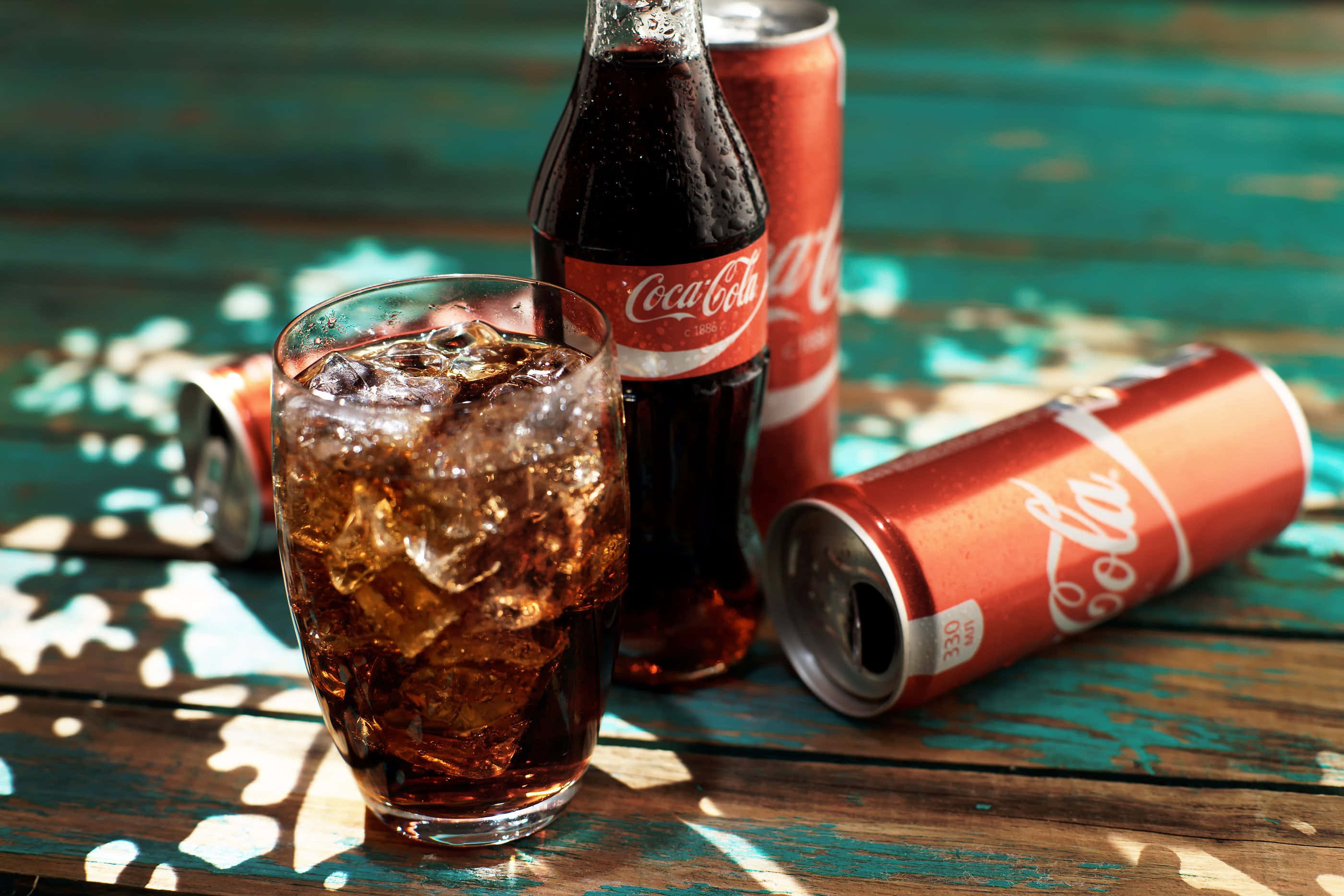 coca cola 桌布 壁紙 — 一杯冰涼的可口可樂放在一個風化的藍綠色木桌上，旁邊有一個瓶子和幾個罐子。
