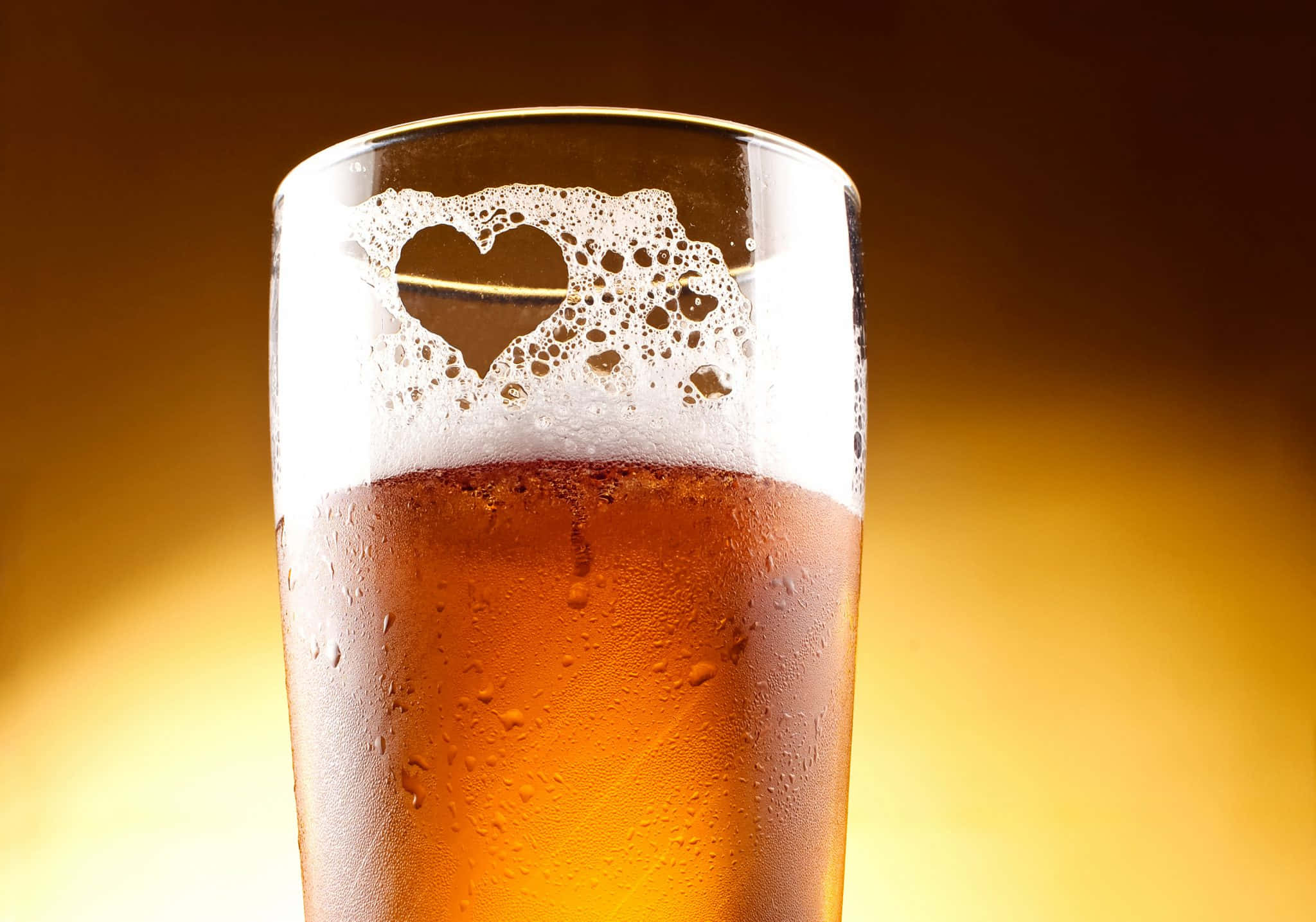 Beer 桌布 壁紙 — 一杯冰涼的琥珀色啤酒，帶有心形泡沫，襯以溫暖的金色漸層背景。