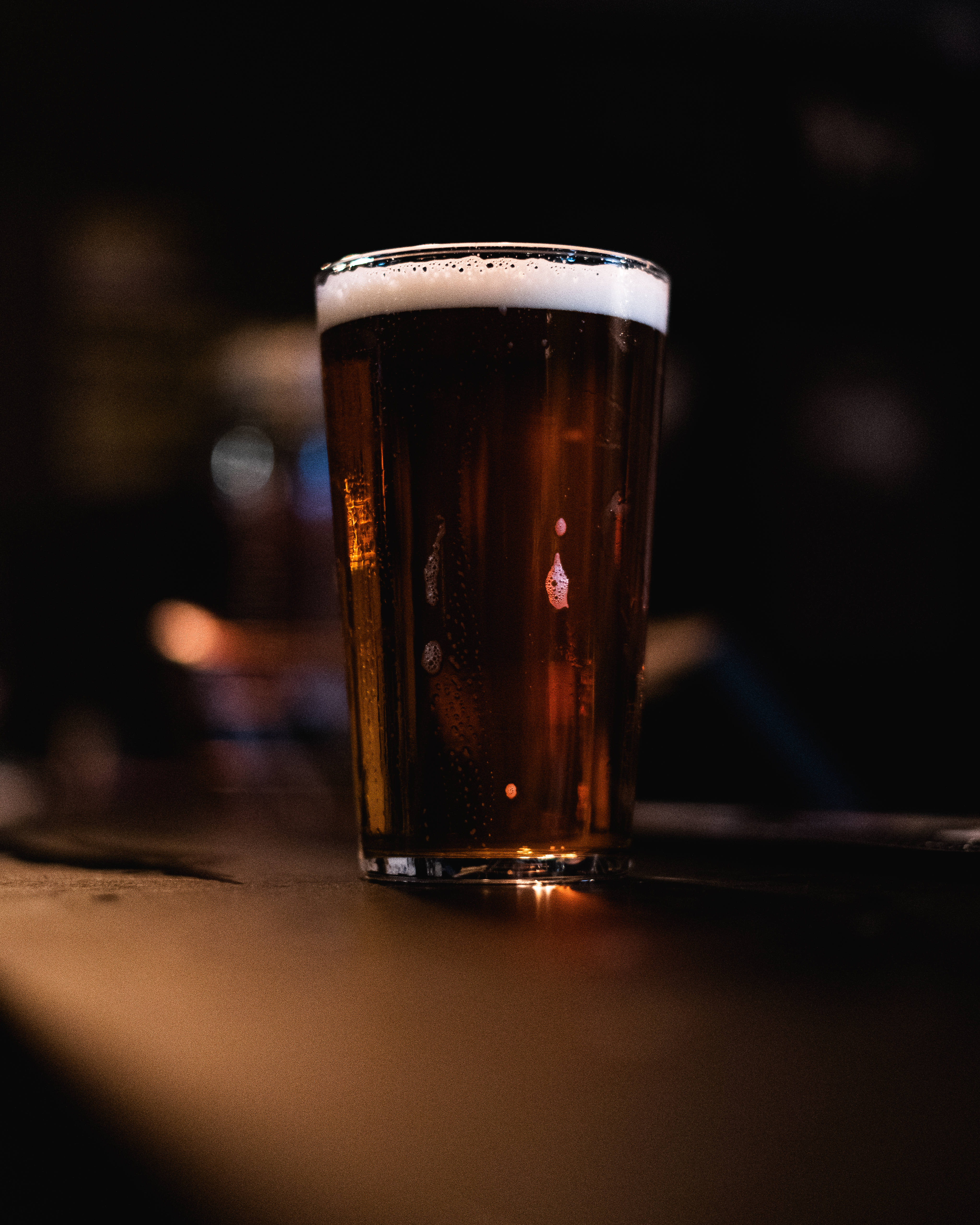 Beer 桌布 壁紙 — 一杯冰涼的琥珀色啤酒，帶有泡沫狀的白色酒頭，放在燈光昏暗的酒吧櫃檯上，營造舒適的酒吧氛圍。