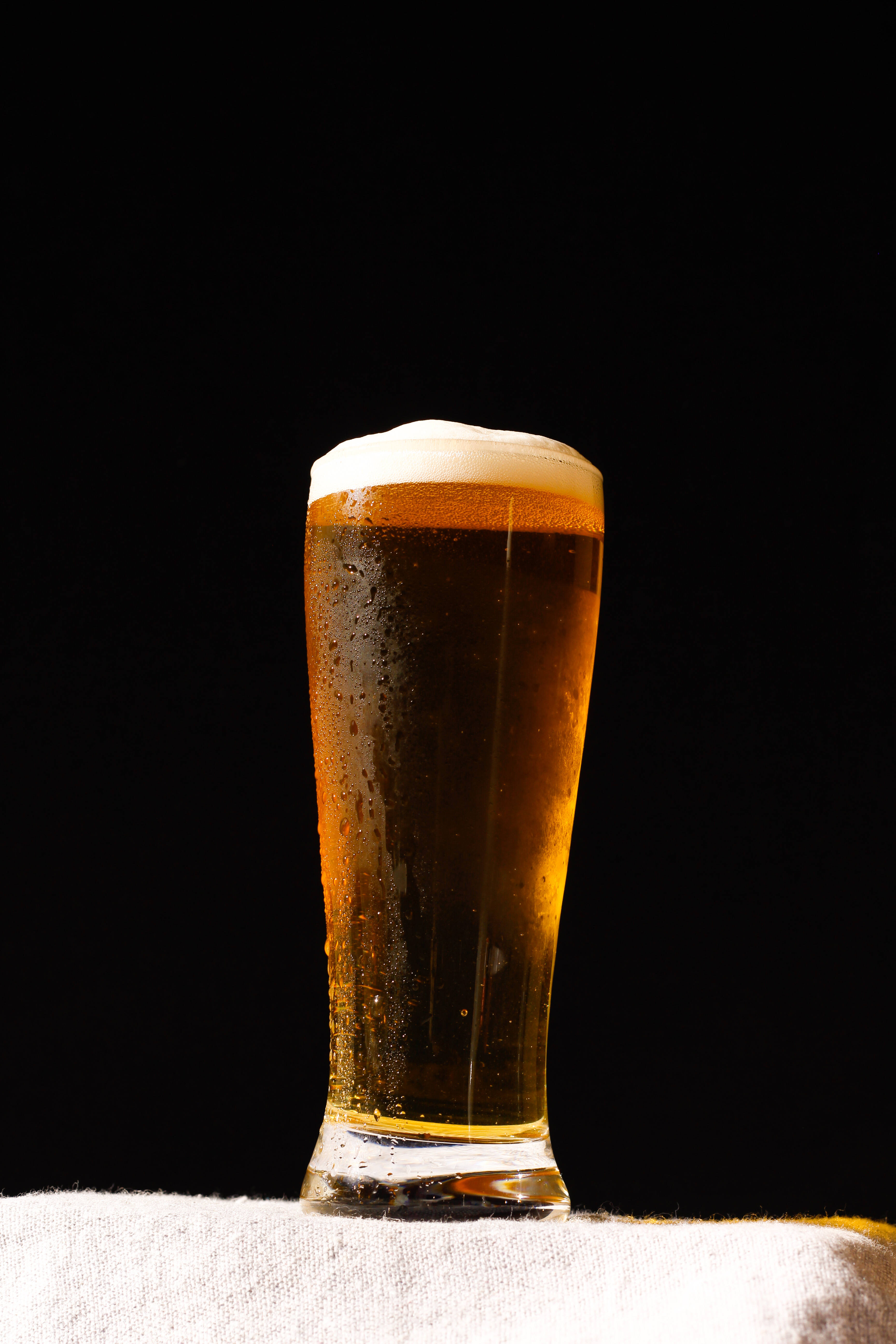Beer 桌布 壁紙 — 一杯冰涼的琥珀色啤酒，帶有白色泡沫，放在白色布上，背景是純黑色。
