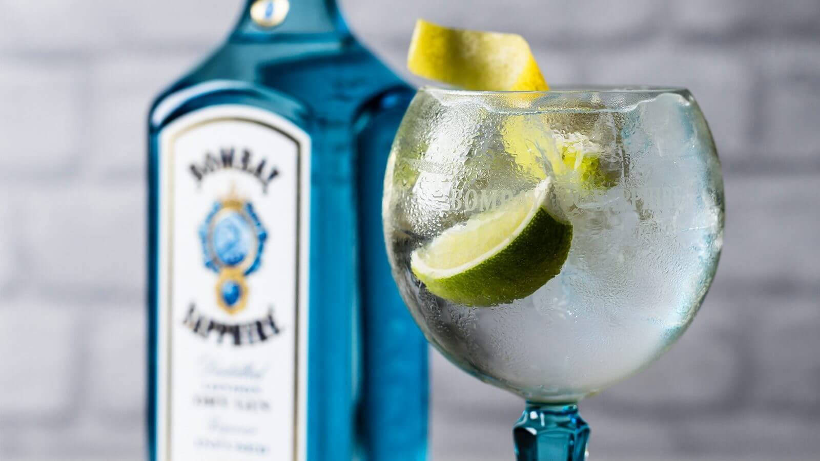 Wine 桌布 壁紙 — 一杯冰涼的琴酒通寧水配青檸角，放在藍色 Bombay Sapphire 酒瓶旁邊，背景是白色磚牆。