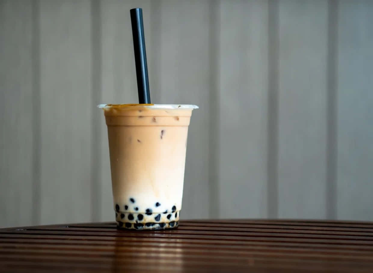 Boba Tea 桌布 壁紙 — 一杯奶油冰珍珠奶茶，內有黑色木薯珍珠，放在透明塑膠杯中，置於木桌上，背景為柔和灰色。