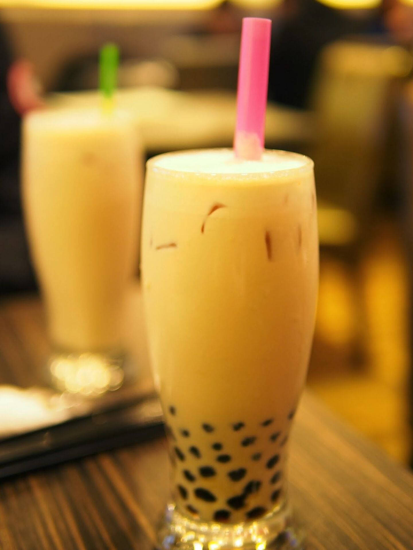 Bubble 桌布 壁紙 — 一杯奶油冰珍珠奶茶，配有黑珍珠和粉紅色吸管，放在溫暖燈光咖啡館的木桌上的一個高身玻璃杯中。