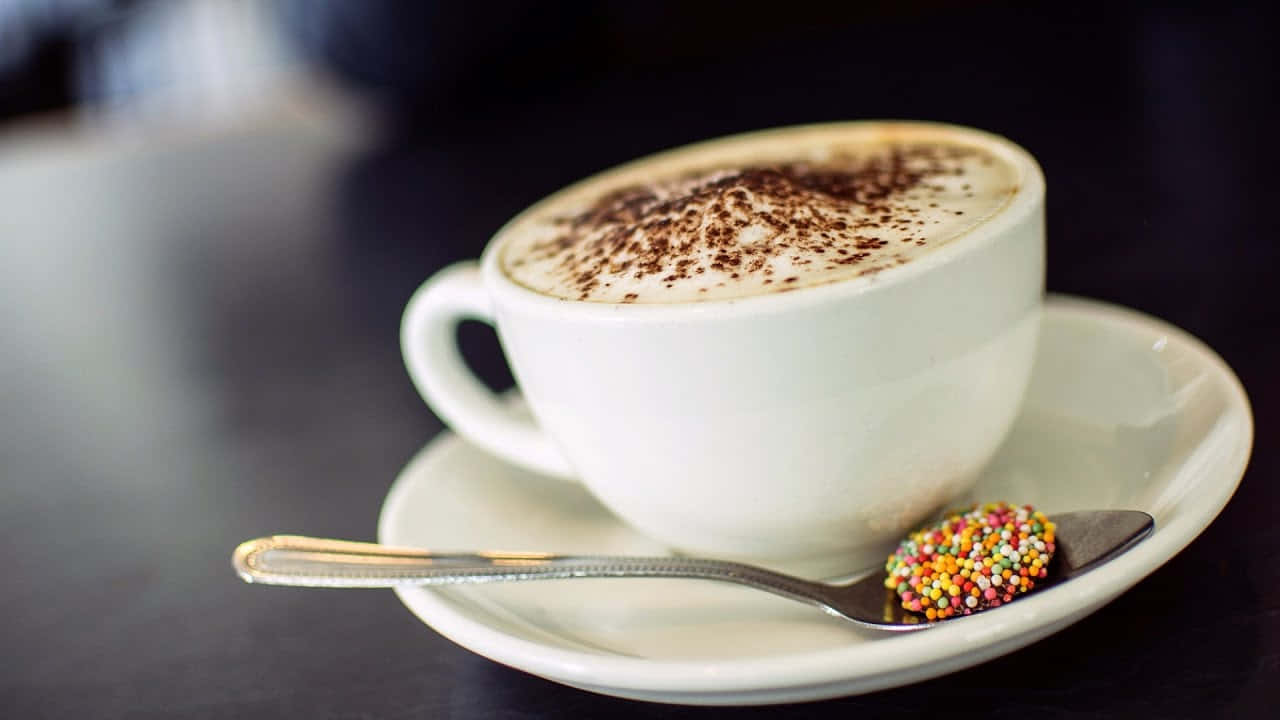Milk And Coffee 桌布 壁紙 — 一杯奶油卡布奇諾，上面撒上可可粉和彩色糖粒，放在碟子上，旁邊有一隻湯匙。
