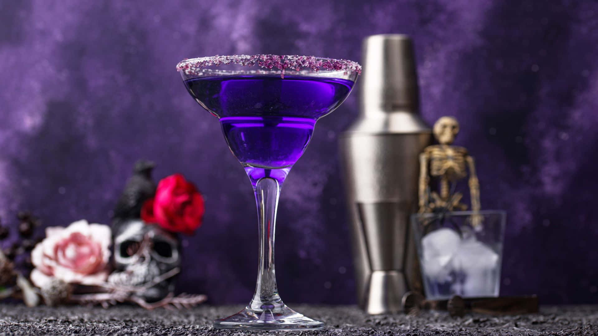 Spooky Cocktails 桌布 壁紙 — 一杯帶有亮粉邊的鮮豔紫色雞尾酒，旁邊放著雞尾酒調酒器和萬聖節主題裝飾，背景為深紫色。