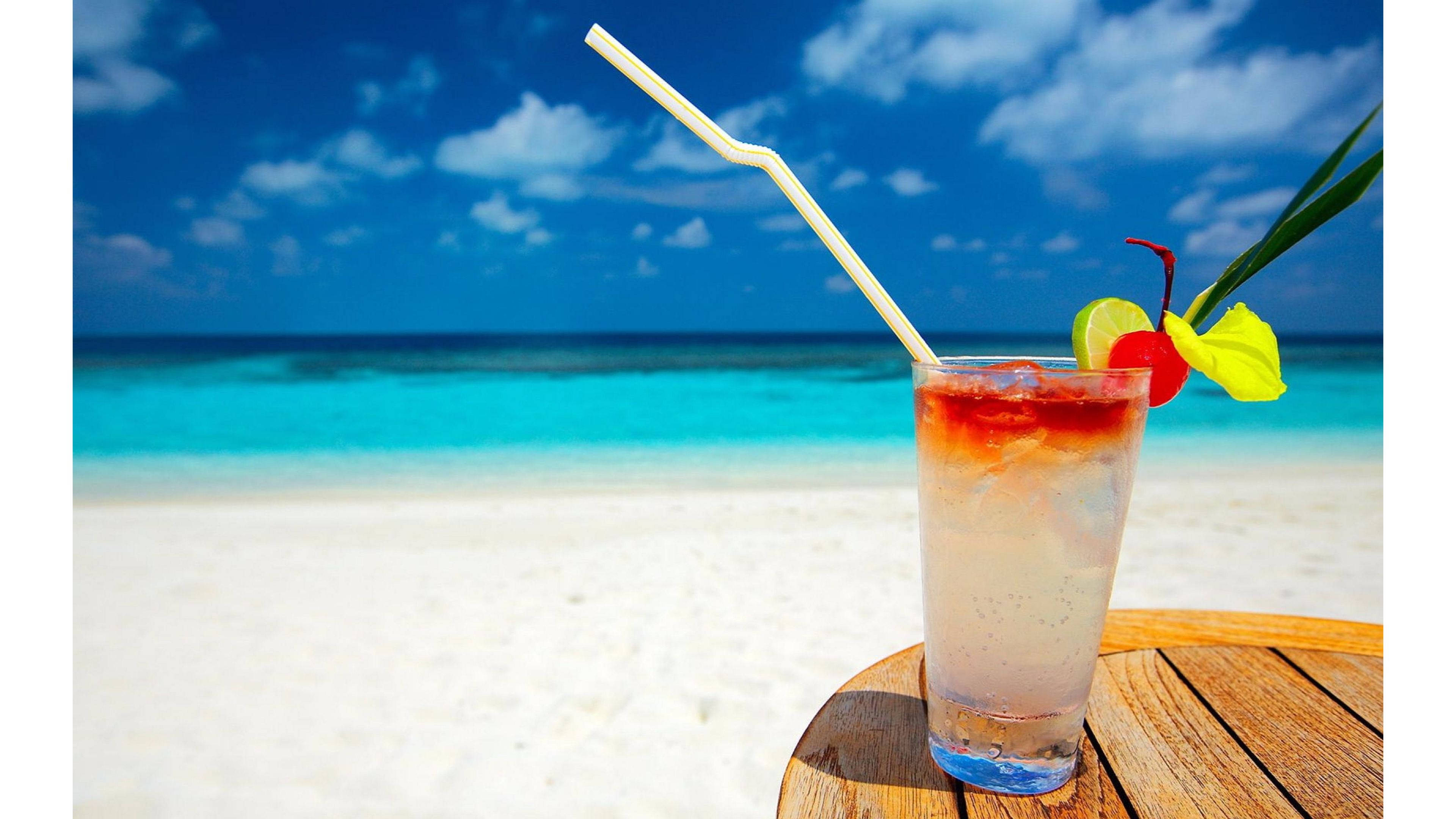 Beach Scenes Desktop 桌布 壁紙 — 一杯帶有吸管和花朵裝飾的熱帶雞尾酒放在木桌上，背景是碧綠的海洋和白色沙灘，上方是明亮的藍天。