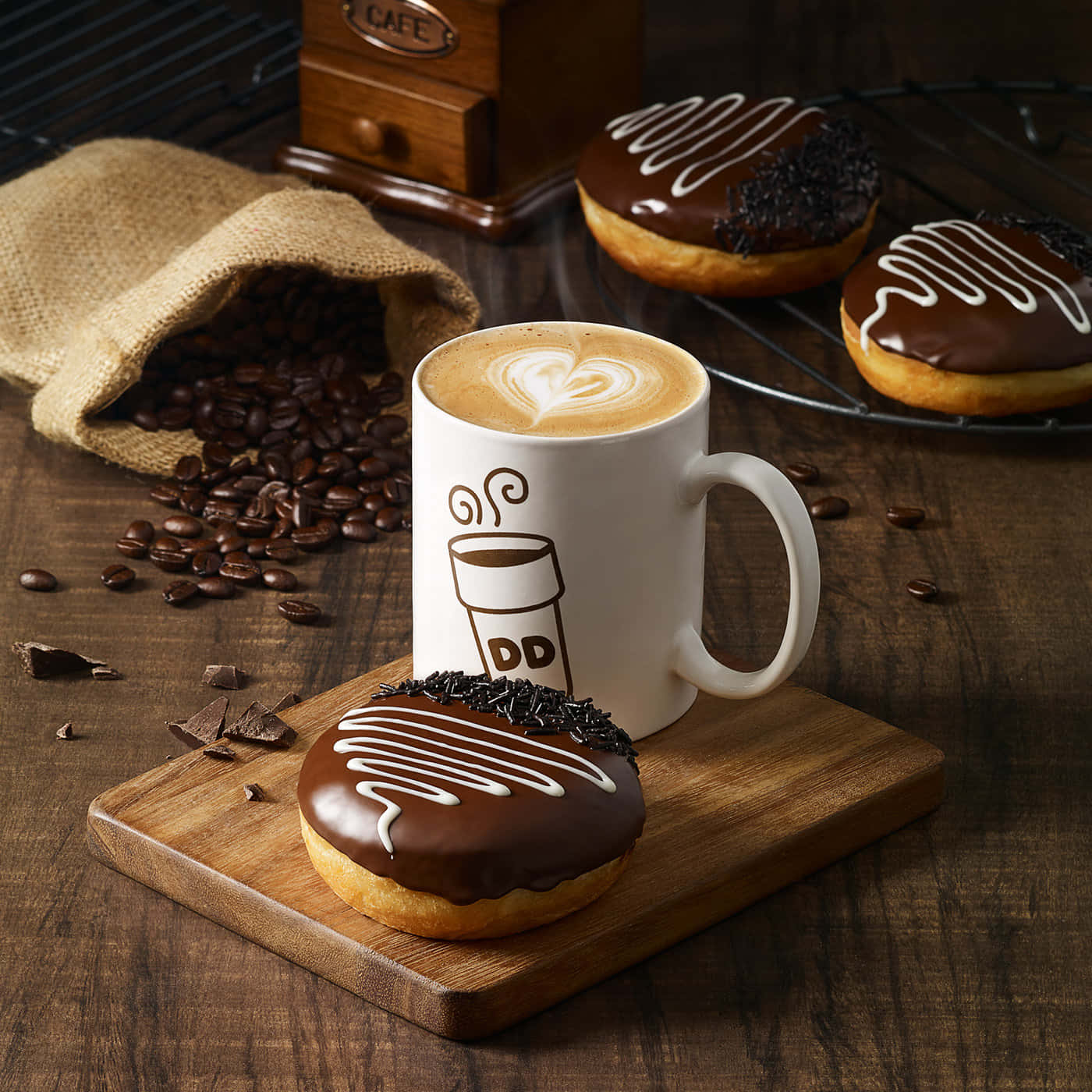 Dunkin Donuts 桌布 壁紙 — 一杯帶有心形拉花的熱拿鐵，旁邊是沾有巧克力的甜甜圈，放在一塊被咖啡豆和麻布袋圍繞的木板上。
