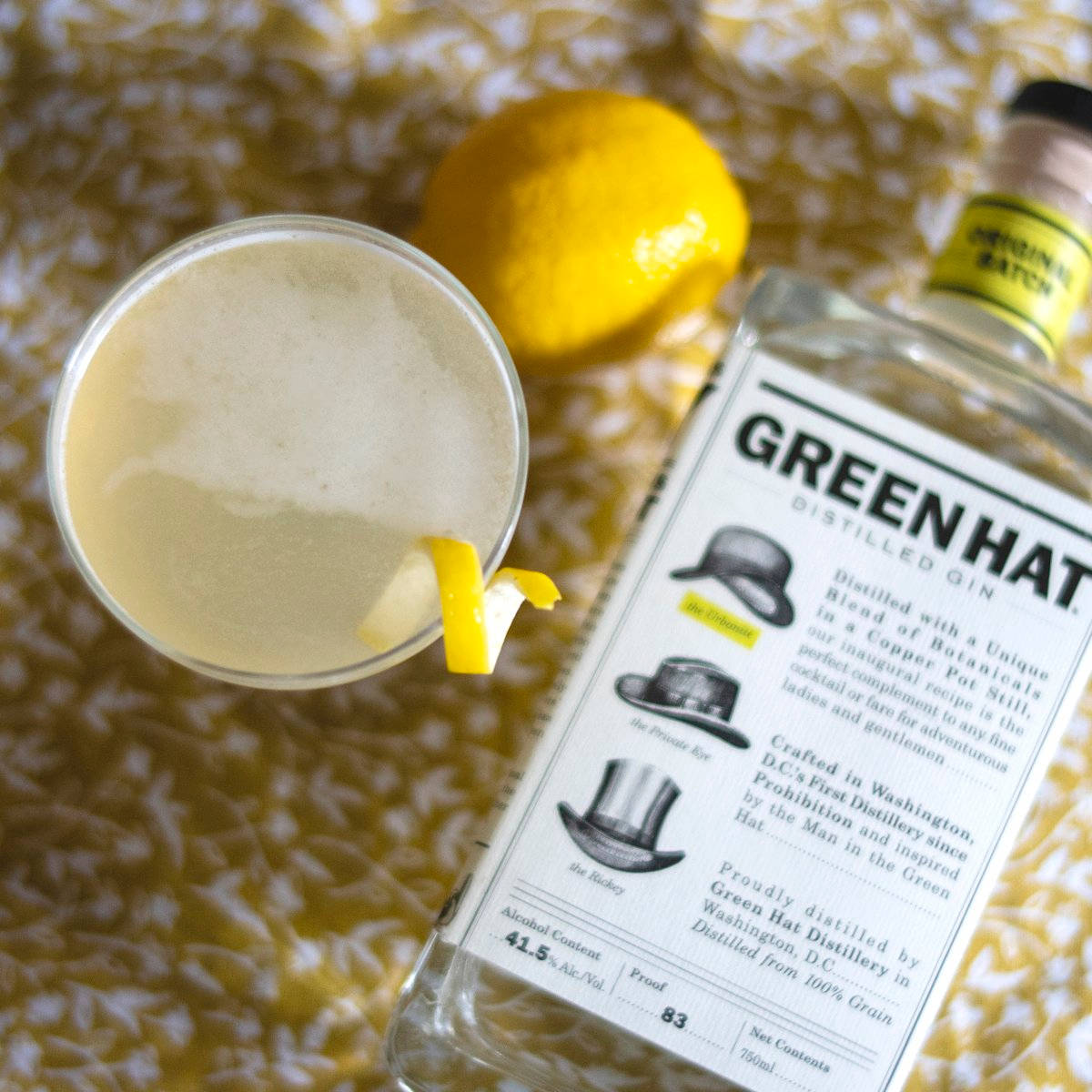 Gin 桌布 壁紙 — 一杯帶有檸檬皮裝飾的泡沫琴酒雞尾酒，旁邊放著一瓶Green Hat琴酒和一個完整的檸檬，置於有紋理的金色表面上。