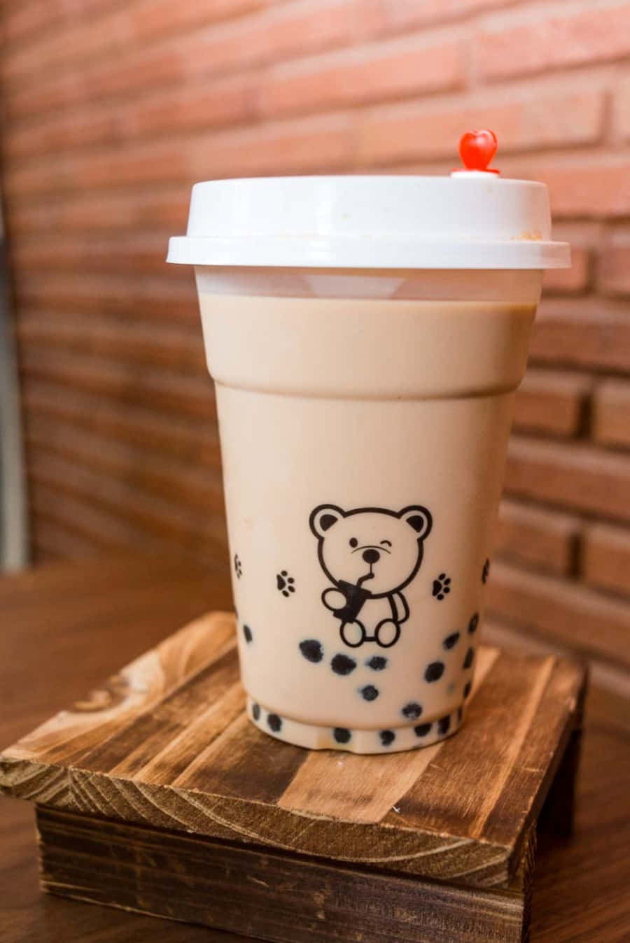 Boba Tea 桌布 壁紙 — 一杯帶有熊設計的白色杯裝奶油泡泡茶，放在木架上，背景是溫暖的磚牆。