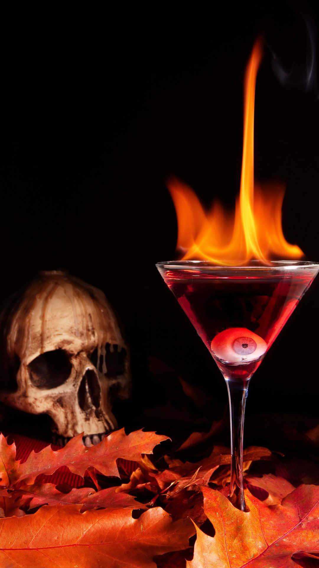 Spooky Cocktails 桌布 壁紙 — 一杯帶有眼球裝飾的火焰紅色雞尾酒放在秋天的樹葉上，旁邊有一個骷髏頭，背景為黑色。