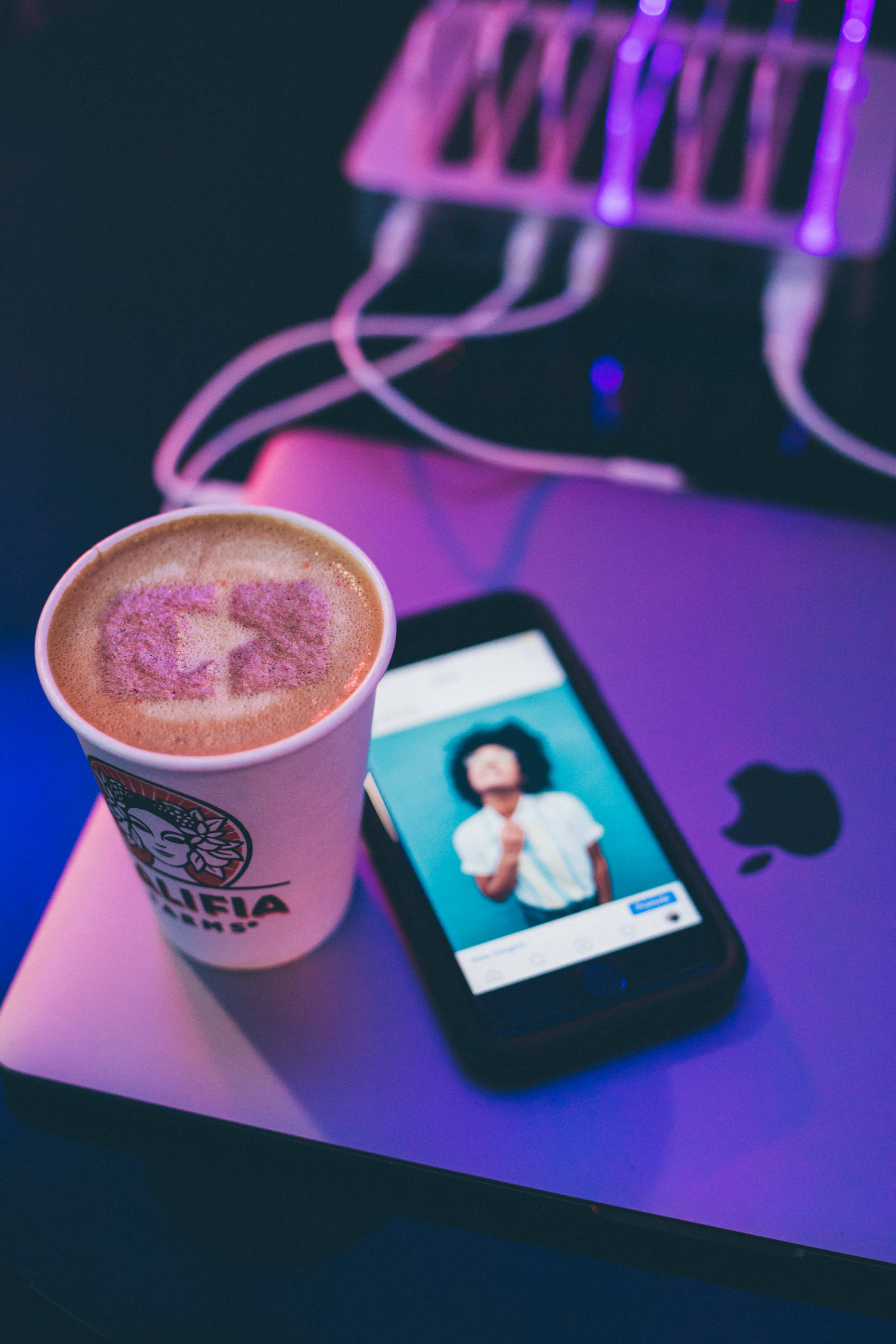 紫色 iPhone 桌布 壁紙 — 一杯帶有粉紅色心形奶泡的拿鐵放在手機旁邊，手機顯示一個人在筆記型電腦上，沐浴在紫色霓虹燈光中。