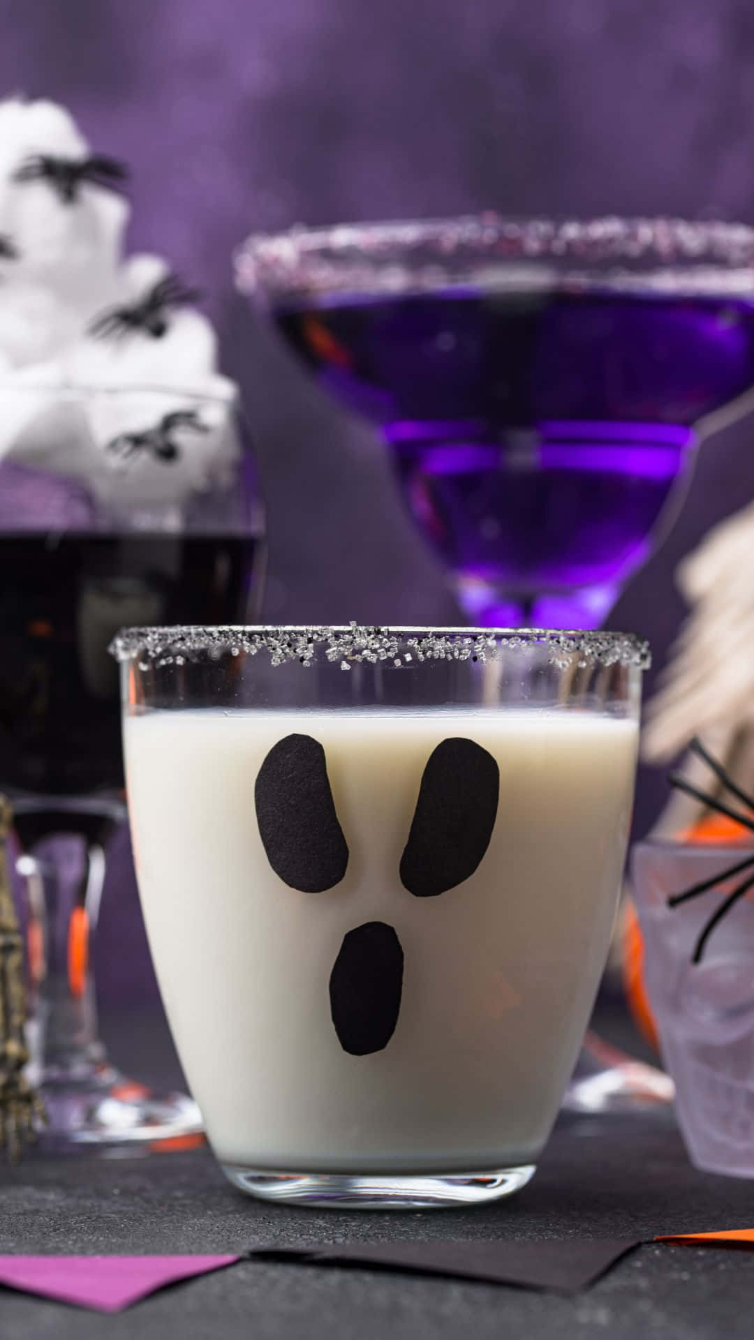 Spooky Cocktails 桌布 壁紙 — 一杯帶有糖邊的幽靈臉奶油雞尾酒，周圍環繞著萬聖節主題飲品和裝飾。