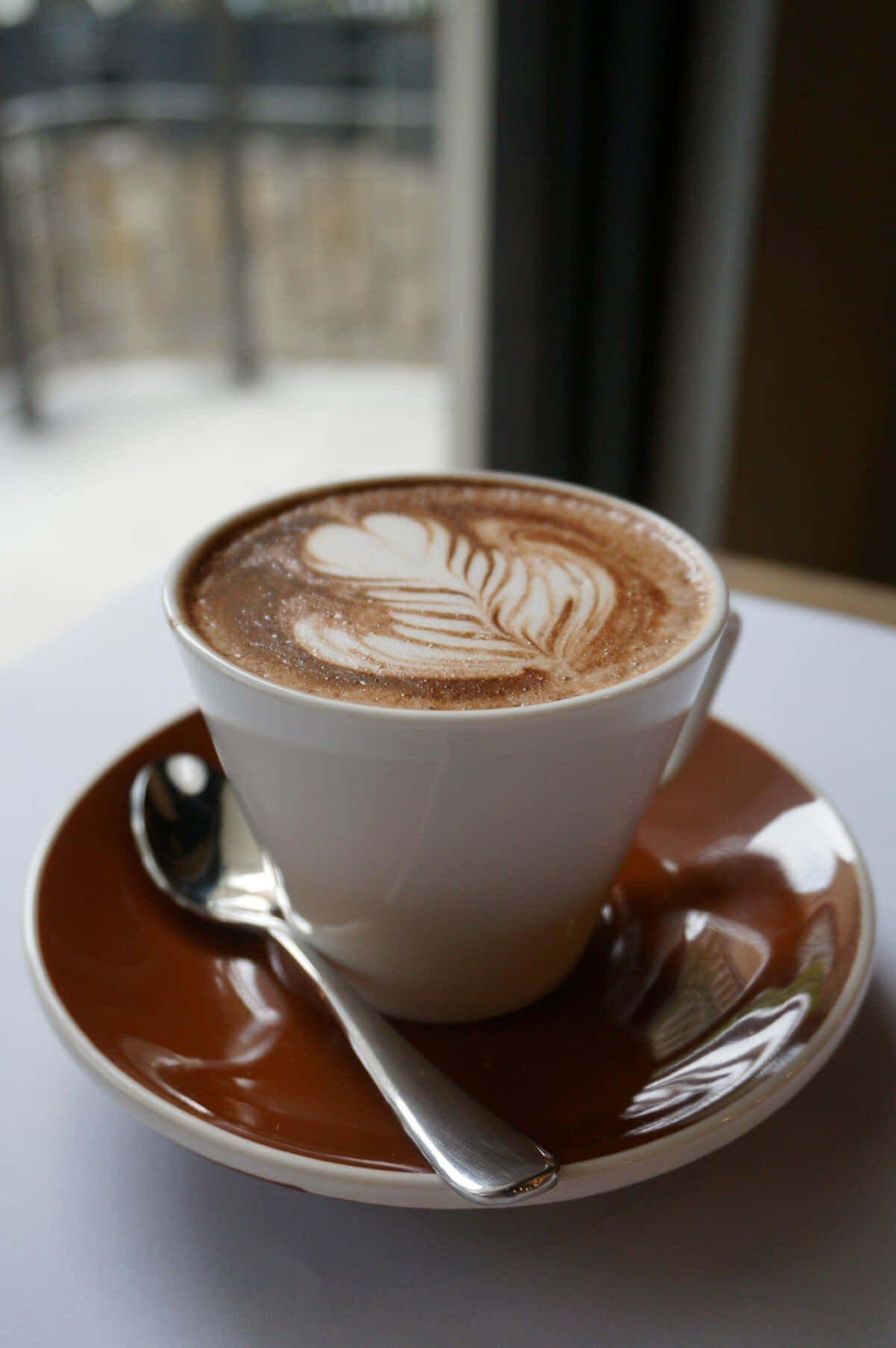 Coffee Cup 桌布 壁紙 — 一杯帶有葉子圖案的白色拉花咖啡，放在棕色碟子上，旁邊有湯匙，靠近窗邊。