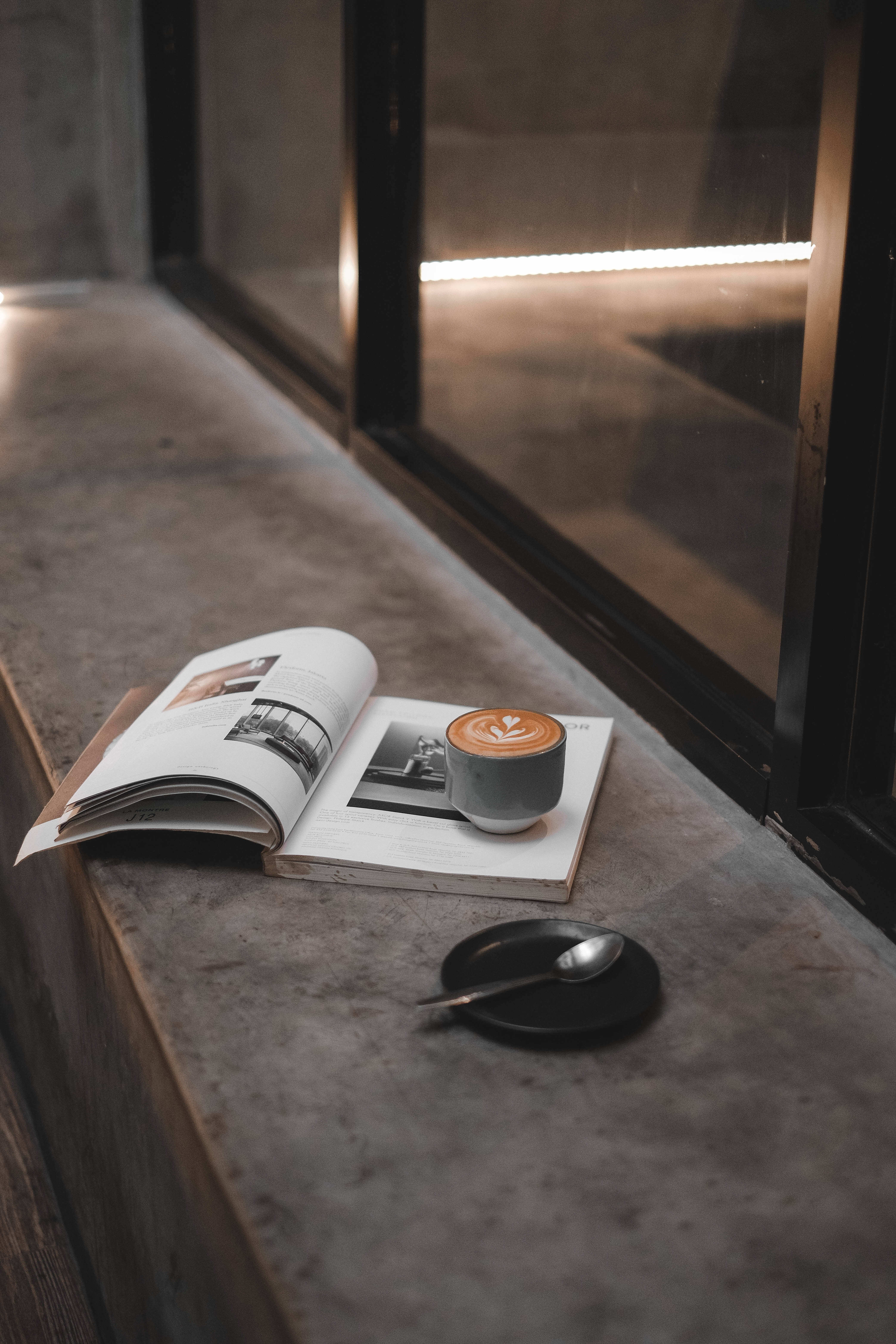 Magazine 桌布 壁紙 — 一杯帶有葉子藝術的拿鐵放在一本打開的雜誌上，旁邊有一隻湯匙在深色碟子上，位於混凝土窗台上，時值黃昏。