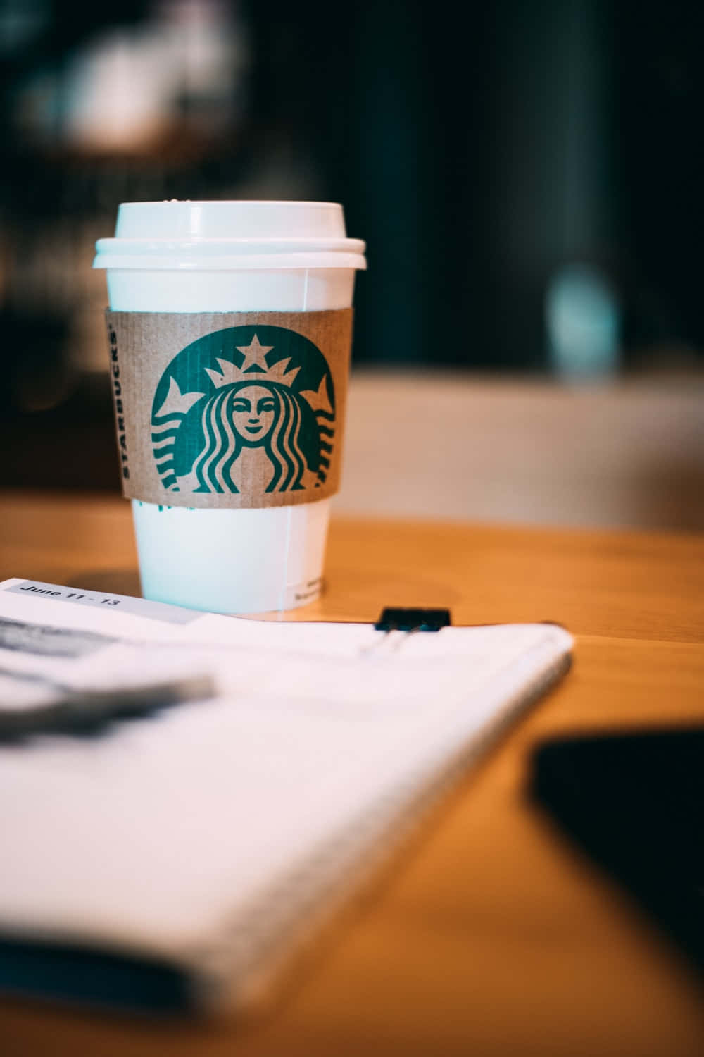 Aesthetic Starbucks 桌布 壁紙 — 一杯星巴克咖啡放在木桌上，旁邊有一本打開的筆記本和一支筆，營造出舒適的咖啡館氛圍。