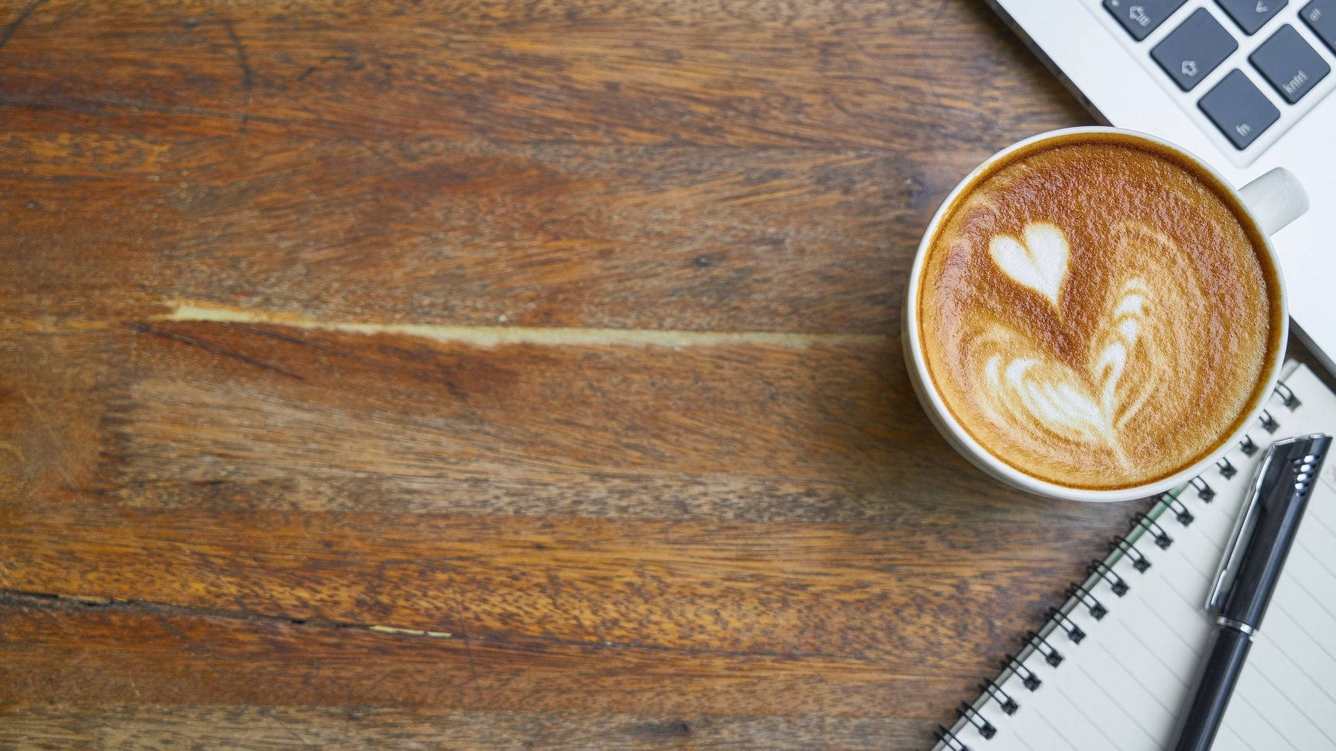 Coffee Aesthetic 桌布 壁紙 — 一杯有心形拉花藝術的咖啡杯放在一個質樸的木桌上，旁邊有筆記型電腦、筆記本和筆。