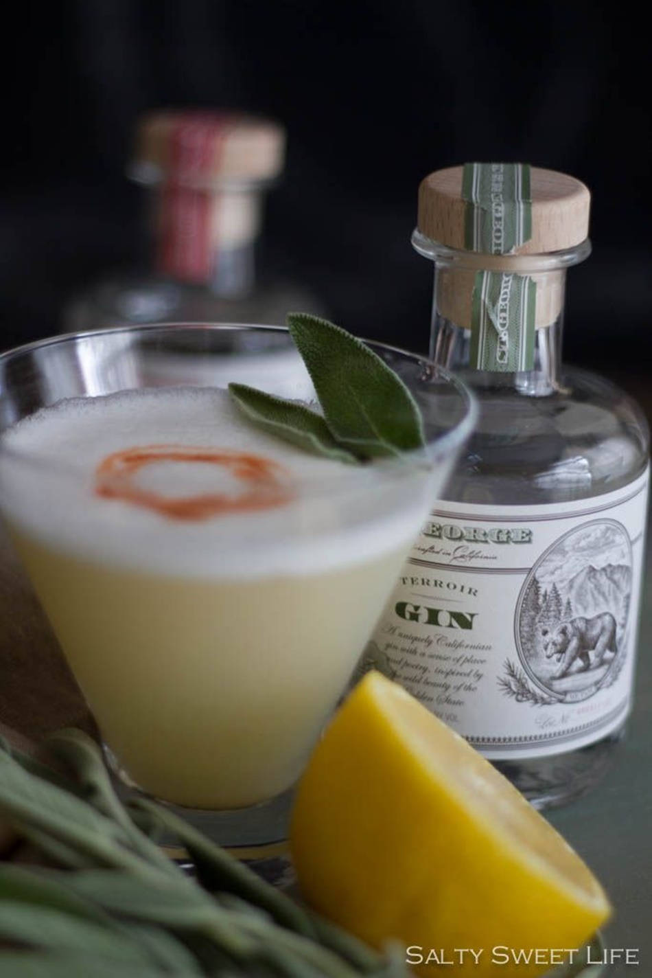 cocktail 桌布 壁紙 — 一杯泡沫琴酒雞尾酒，用鼠尾草和檸檬角裝飾，旁邊放著一瓶Terroir Gin，背景深色。
