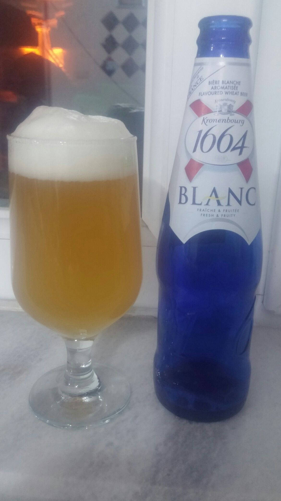 Beer Bottle 桌布 壁紙 — 一杯泡沫豐富的Kronenbourg 1664 Blanc啤酒放在其藍色瓶子旁邊，位於溫暖壁爐附近的大理石檯面上。