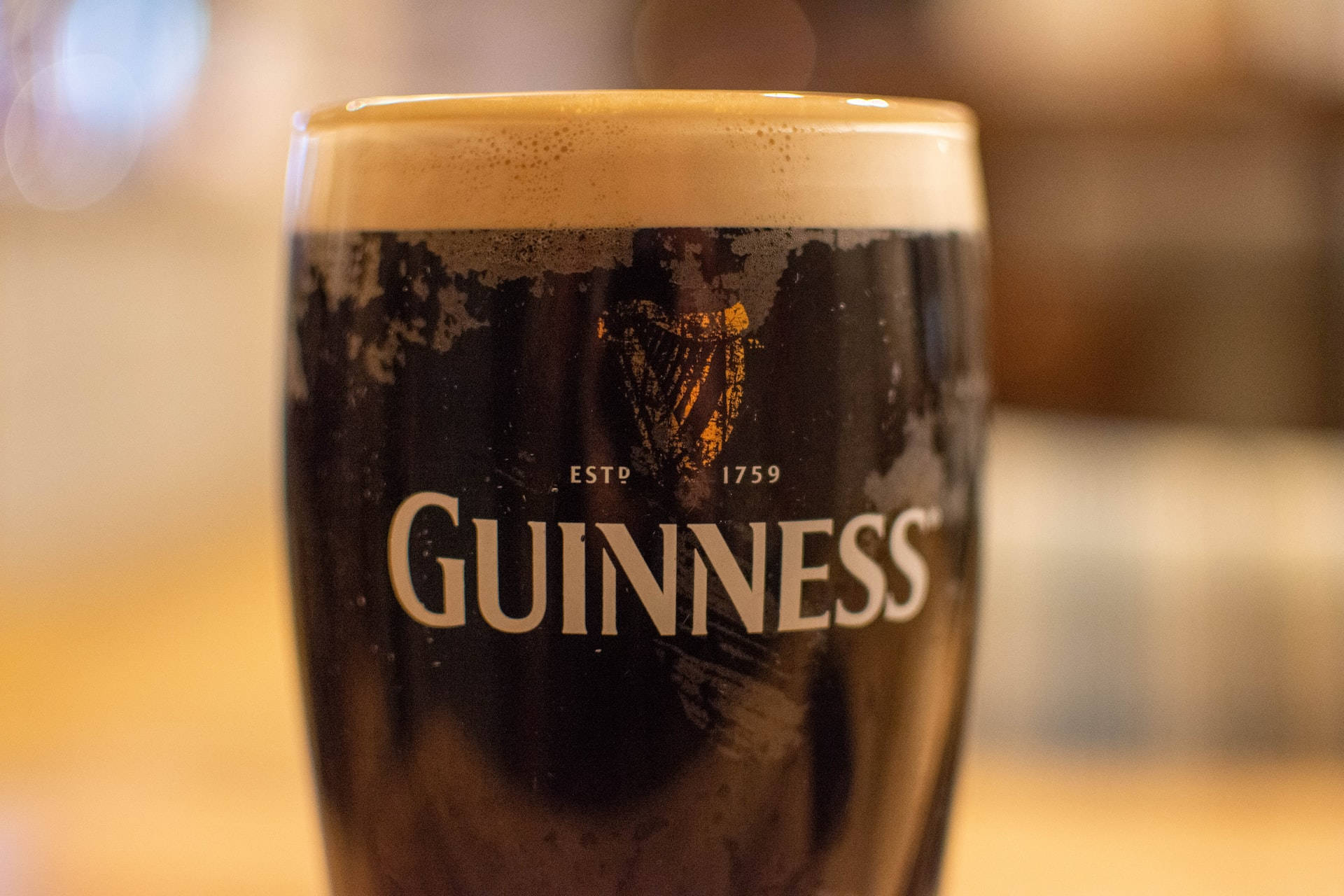 Beer 桌布 壁紙 — 一杯泡沫豐富的健力士啤酒特寫，帶有金色泡沫和標誌性的豎琴標誌，背景是溫暖模糊的酒吧。