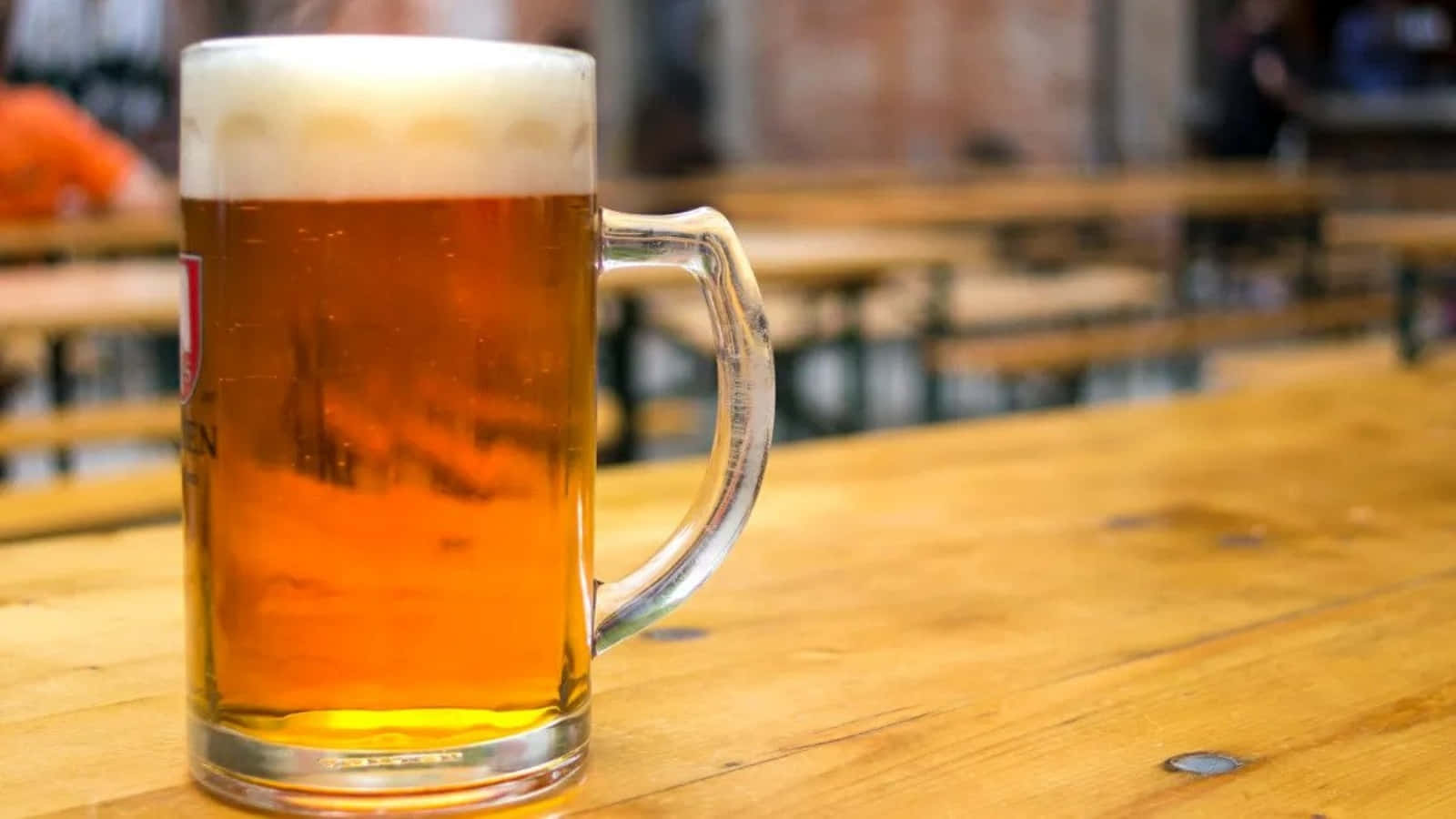 Beer 桌布 壁紙 — 一杯泡沫豐富的琥珀色啤酒放在質樸的木桌上，背景是模糊的長凳，營造出舒適的啤酒館氛圍。