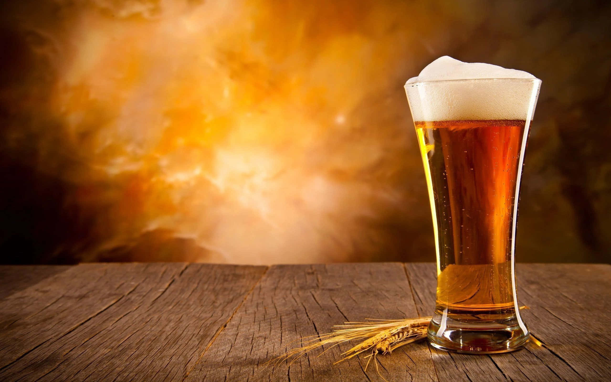 Beer 桌布 壁紙 — 一杯泡沫豐富的琥珀色啤酒放在高身玻璃杯中，置於鄉村風格的木桌上，旁邊有小麥莖，背景溫暖發光。