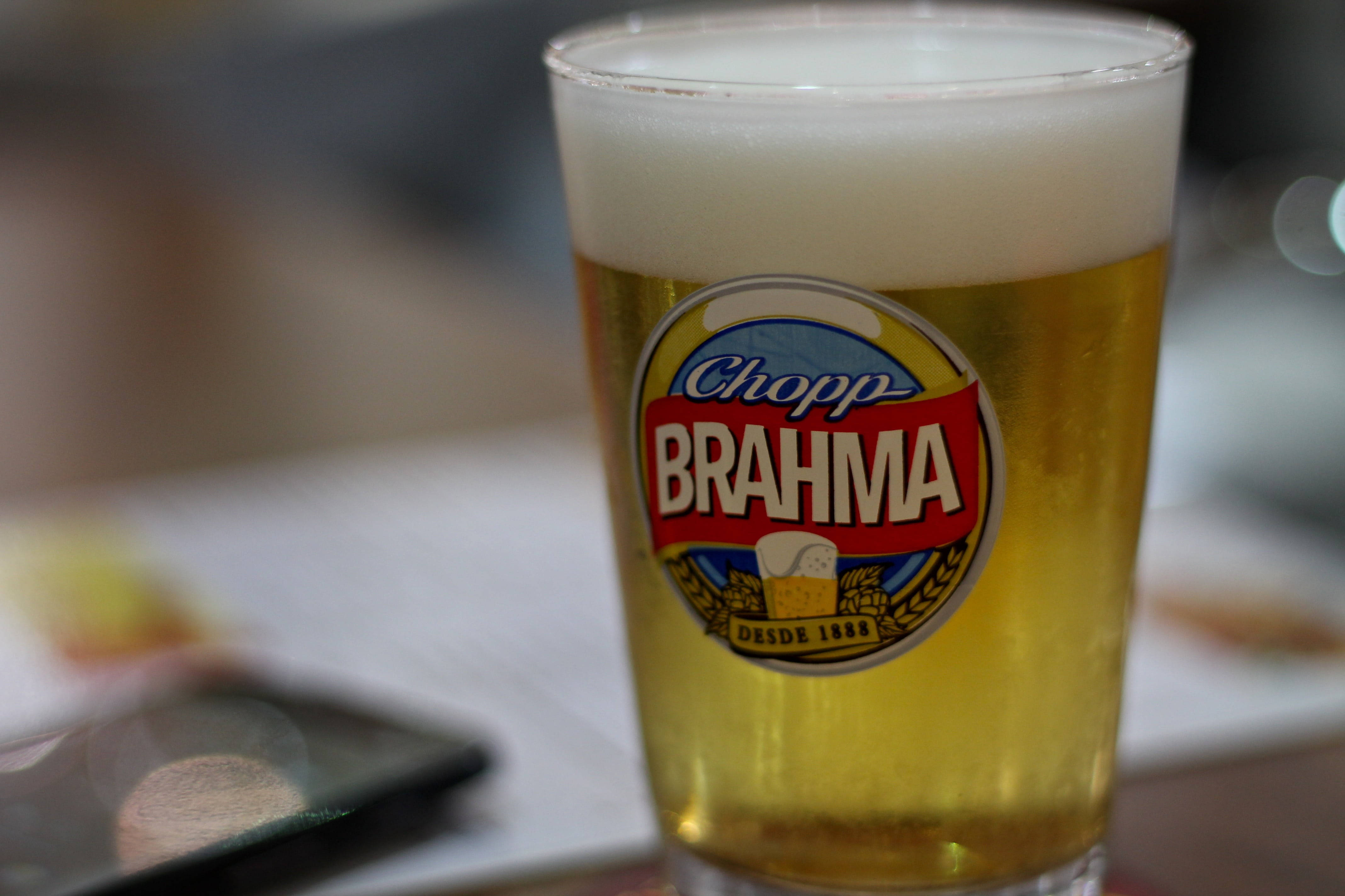 Drink 桌布 壁紙 — 一杯泡沫豐富的金色Brahma啤酒，帶有醒目標誌，放在桌上，背景是模糊的電話。