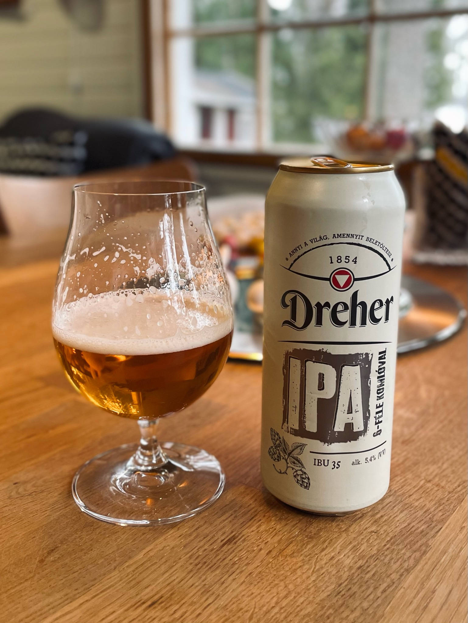 Beer 桌布 壁紙 — 一杯泡沫豐富的金色IPA啤酒放在鬱金香杯中，旁邊放著它的罐頭，在窗邊溫暖的木桌上。