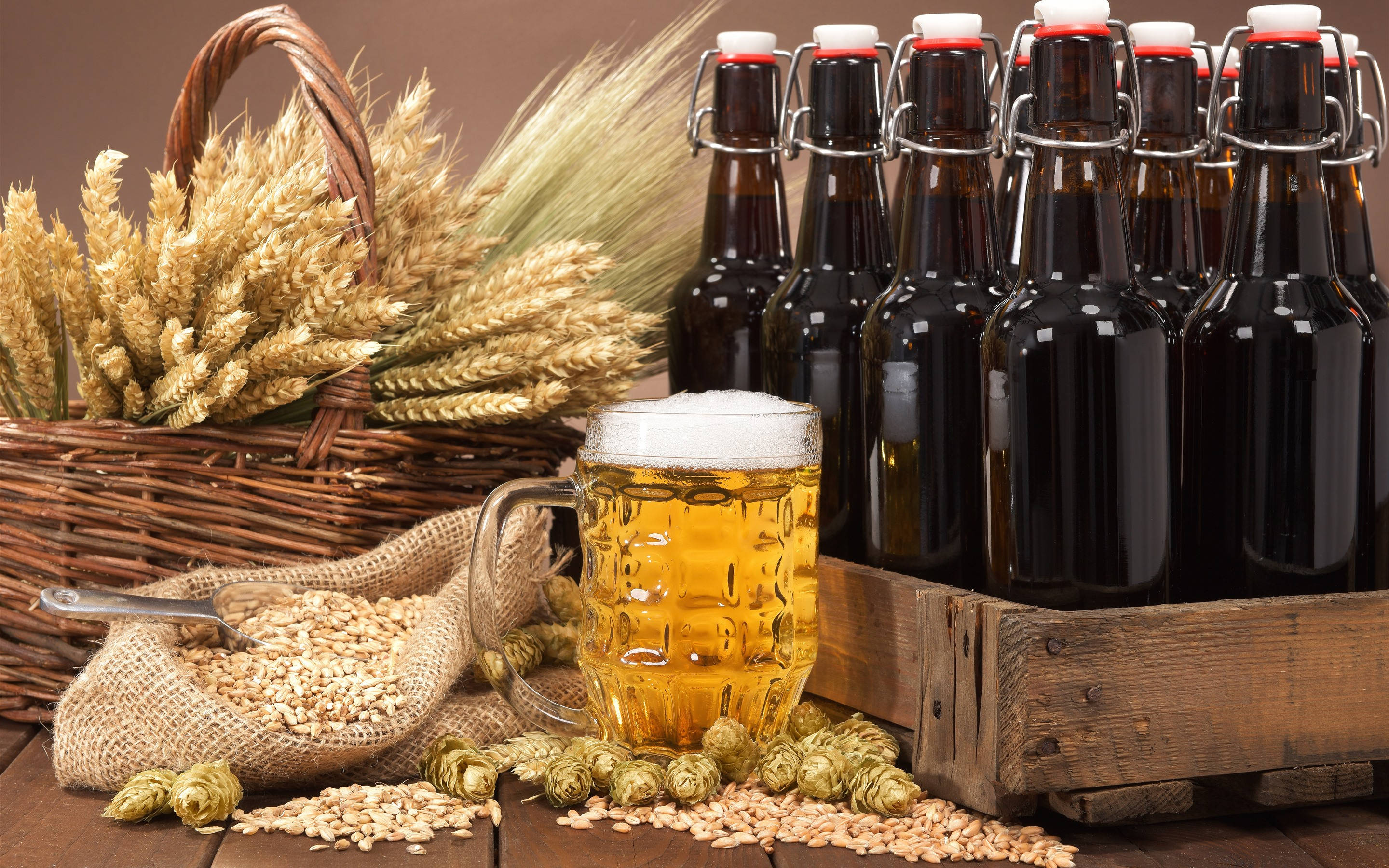 alcohol 桌布 壁紙 — 一杯泡沫豐富的金色啤酒放在一個木箱旁邊，木箱裡有深色瓶子和一籃小麥，擺在質樸的木桌上。