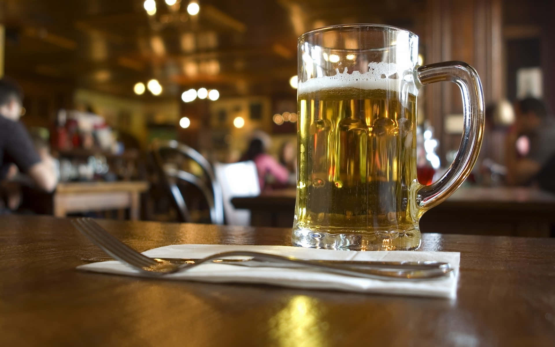 Beer 桌布 壁紙 — 一杯泡沫金黃啤酒放在溫暖燈光的酒吧木桌上，背景有模糊的顧客。