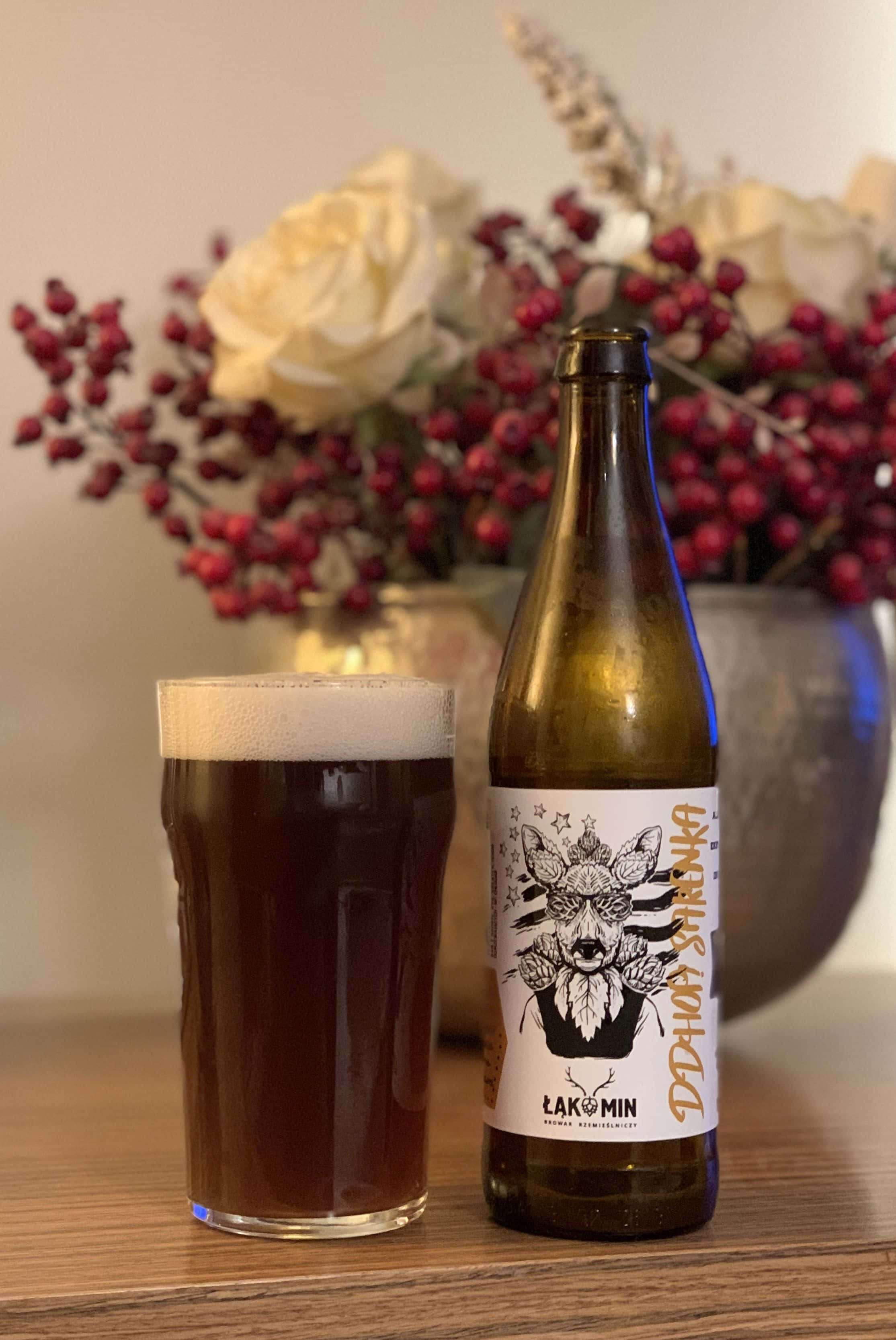 Beer 桌布 壁紙 — 一杯深琥珀色啤酒帶有泡沫，旁邊放著標有 'Lakomin' 的瓶子，在木桌上，背景柔和，有一瓶白玫瑰和紅莓。